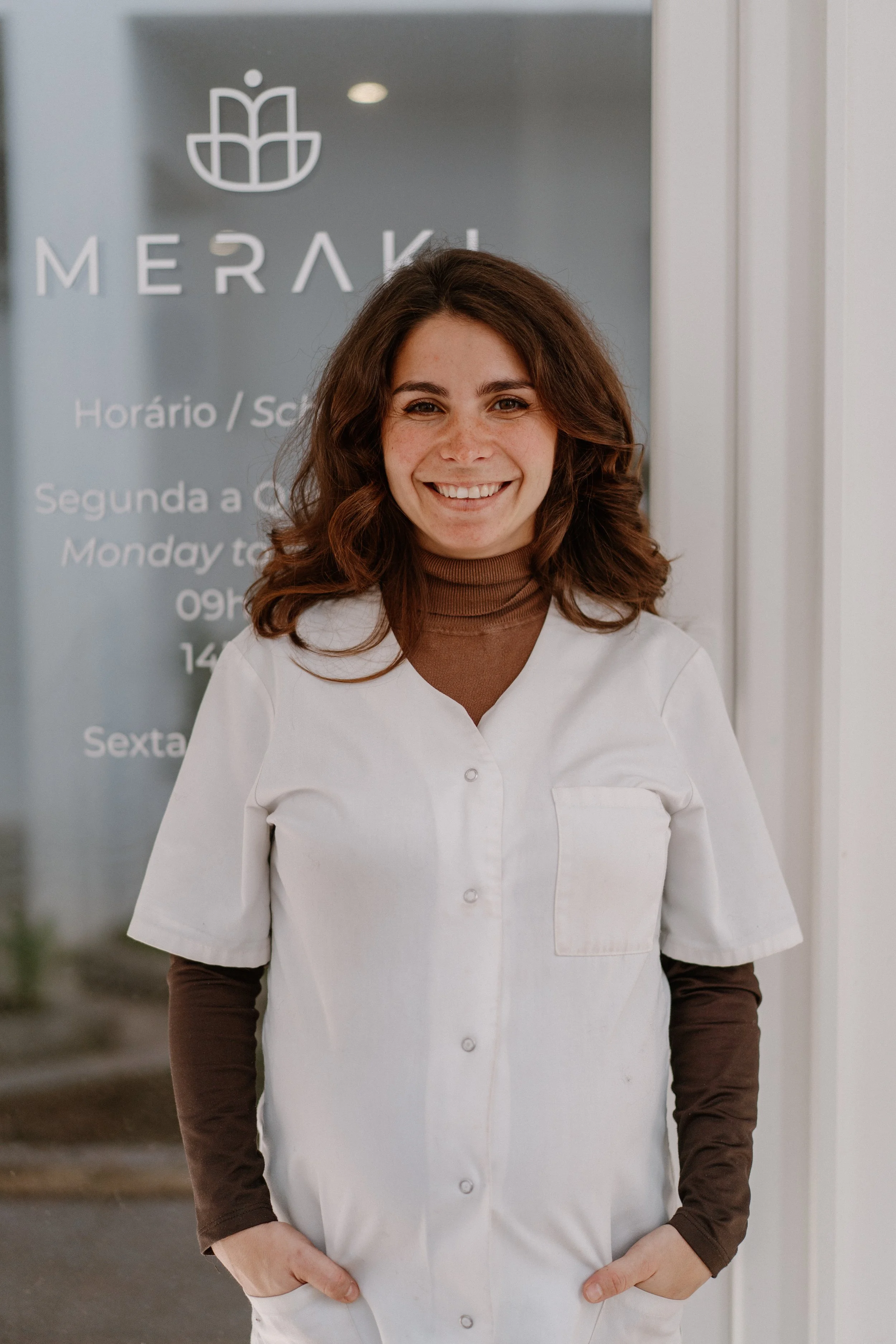 A young woman with brown hair, wearing a brown turtleneck and a white medical coat with her hands in her pockets, standing in front of a grey background with a logo and text.