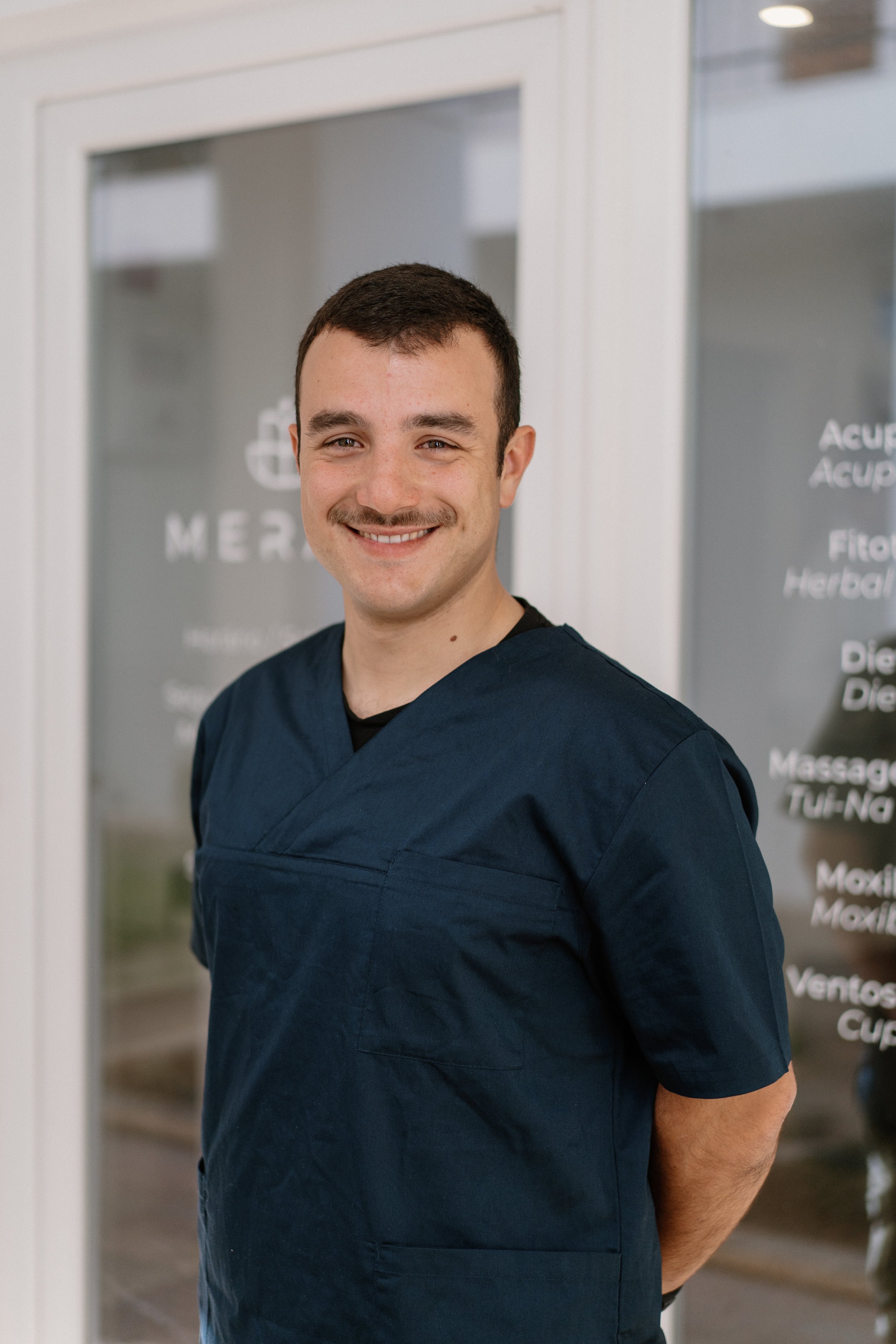 A smiling young man with short dark hair and a mustache wearing dark navy scrubs, standing indoors with a medical office or clinic background.