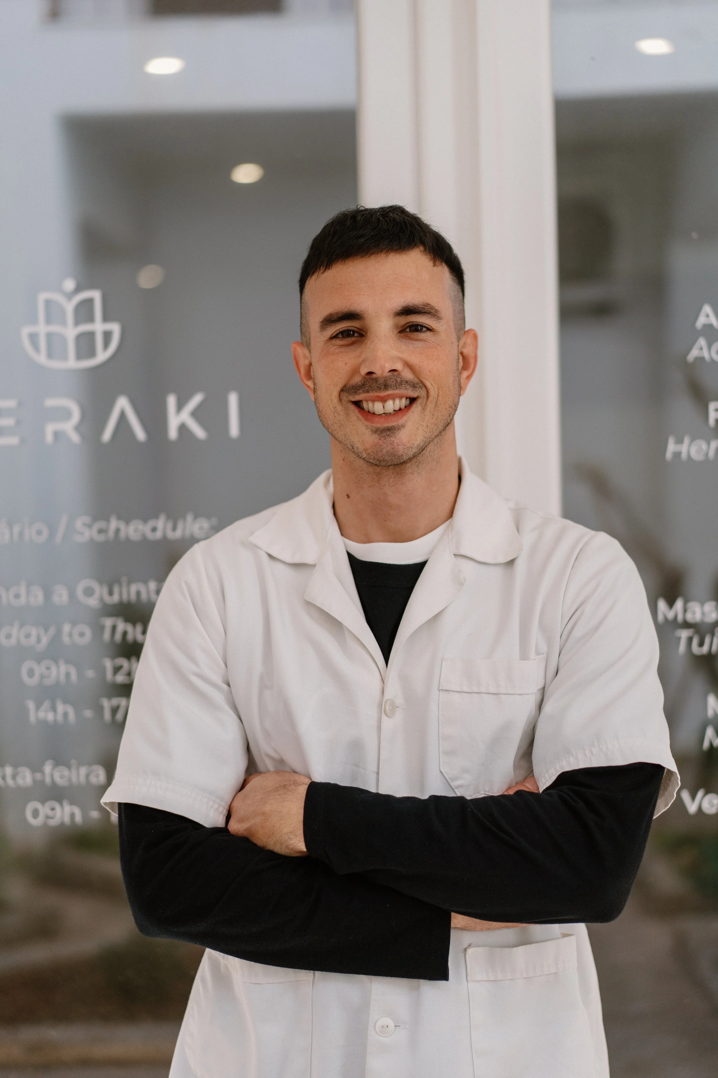 A smiling man with short dark hair, wearing a white coat over a black long-sleeve shirt, standing with arms crossed in front of a glass window with text and a logo.
