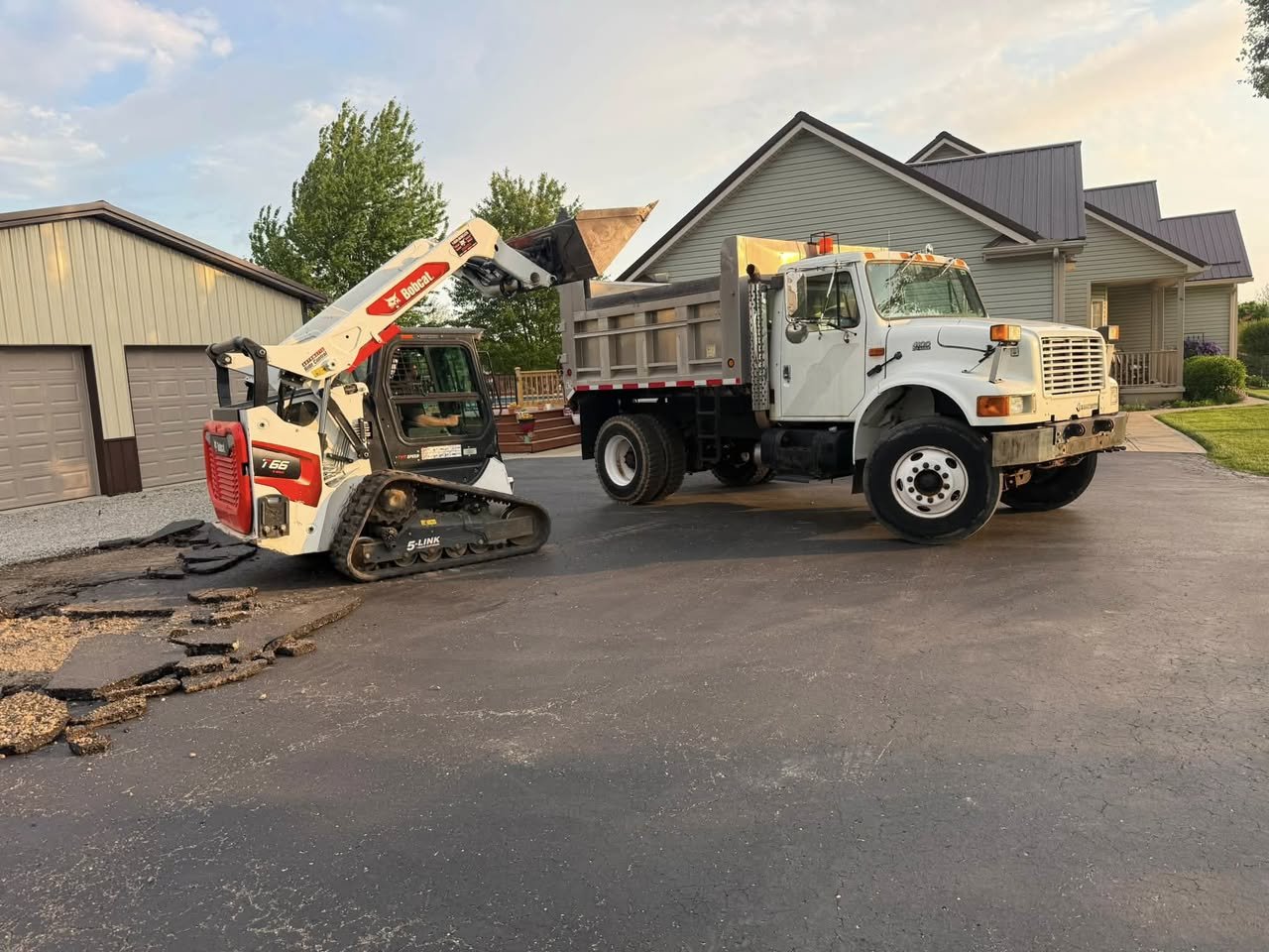 A small bobcat skid-steer loader lifting debris into the bed of a large white dump truck on a residential driveway during daylight hours.