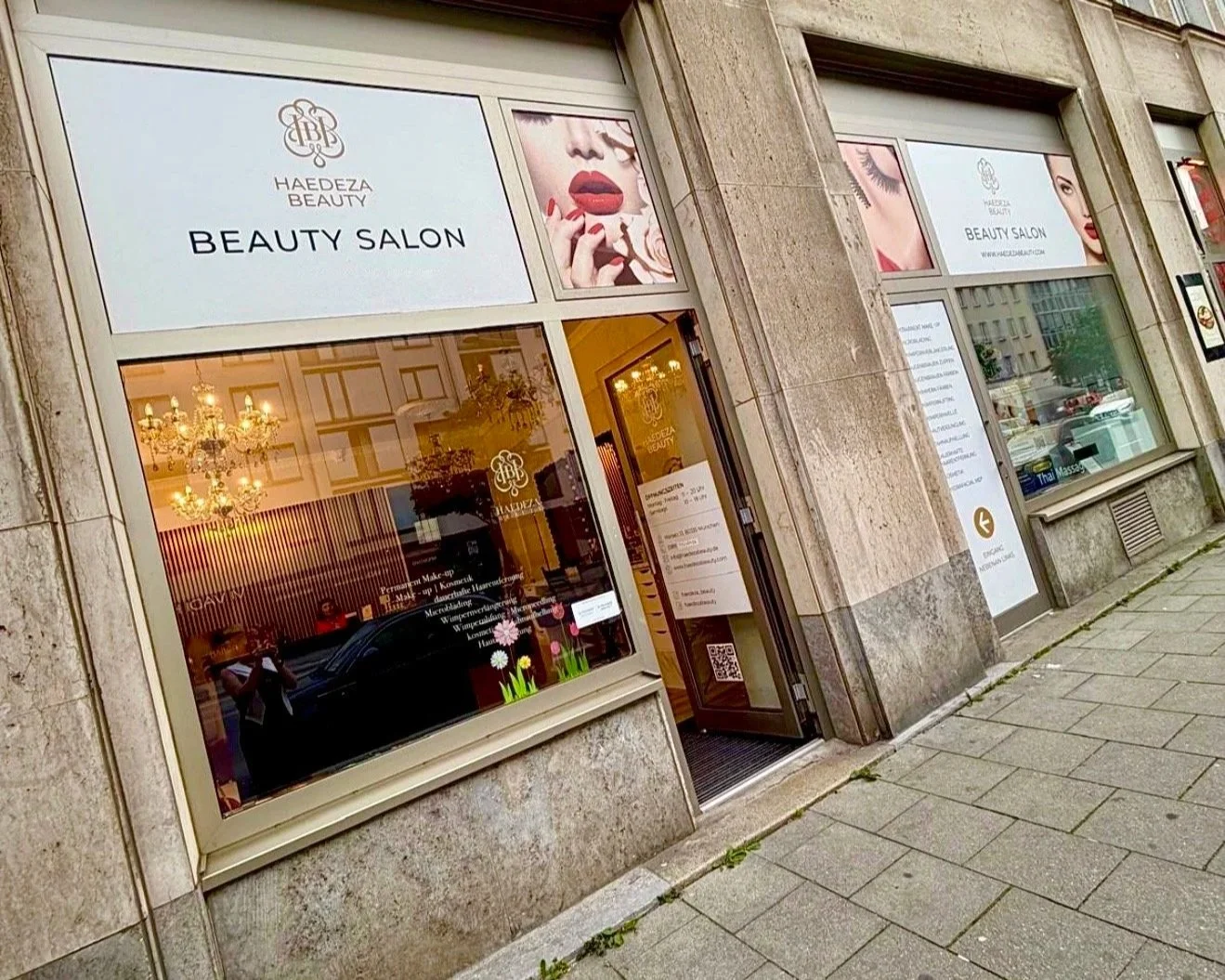 Außenansicht eines Schönheitssalons mit Schaufenster und Eingang in einem Gebäude mit Steinfassade, Schaufenster zeigt Dekorationen und Chandelier, Werbung für Beauty-Behandlungen, teilweise reflektiert.