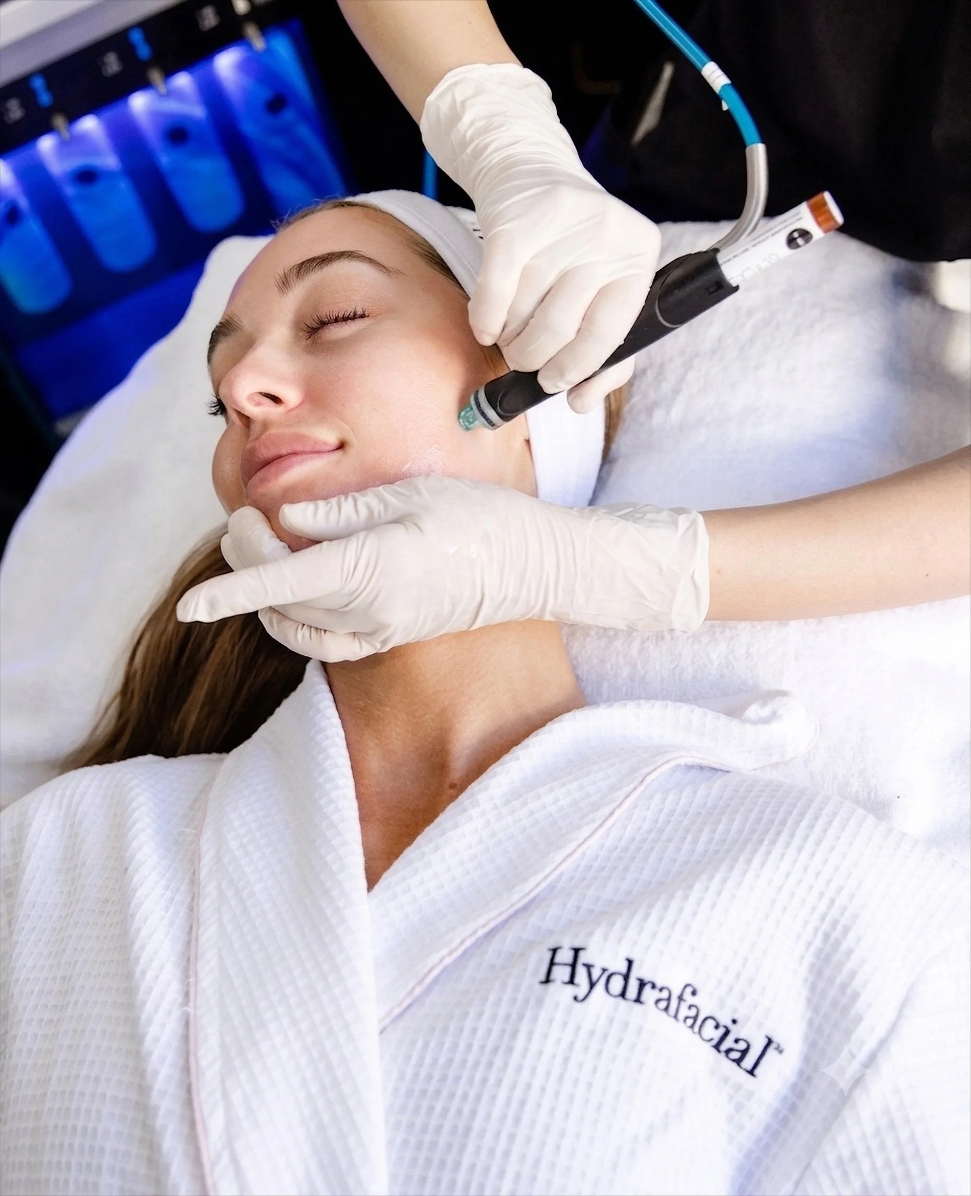 Eine Frau liegt entspannt im weißen Arztkittel mit der Aufschrift 'Hydratisierung'. Ein Arzt führt eine kosmetische Laserbehandlung im Gesicht durch, trägt medizinische Handschuhe und verwendet ein medizinisches Gerät.