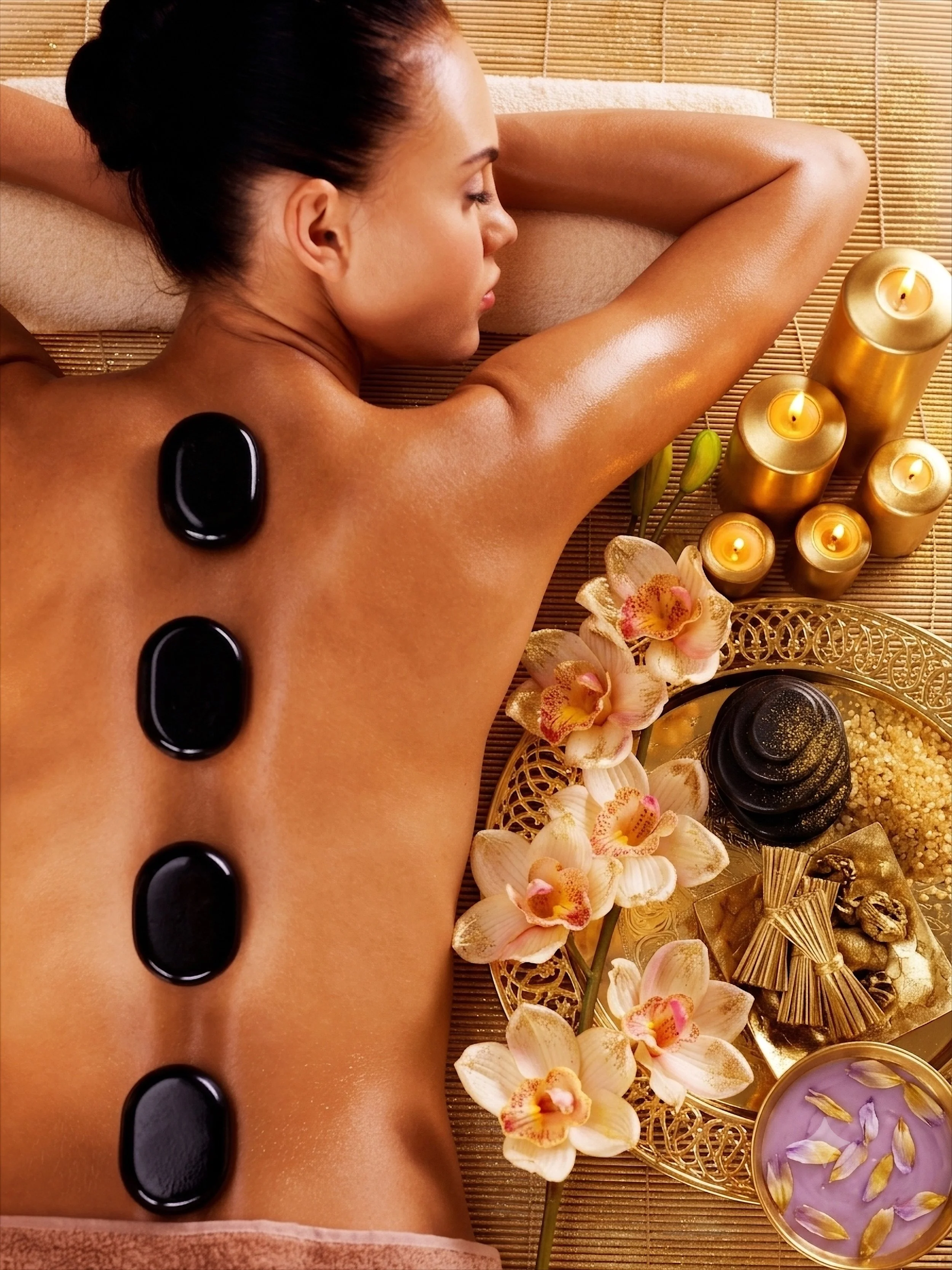 Frau liegt entspannt auf einer Massagebank, umgeben von Kerzen, Orchideen und Spa-Accessoires, während Steine auf ihrem Rücken liegen.