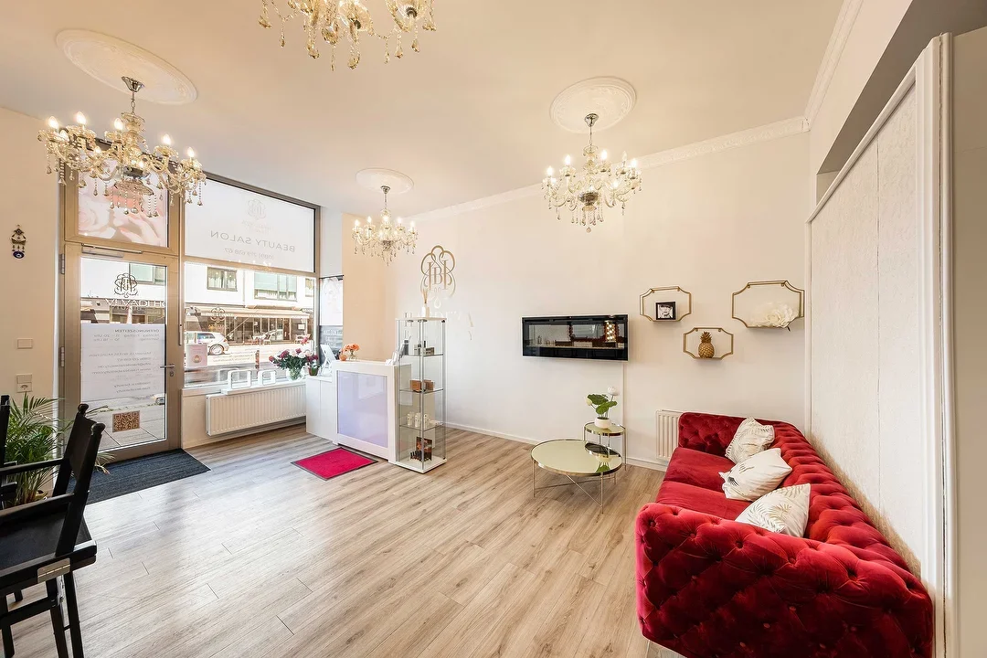 Innenraum eines eleganten Salons mit weißen Wänden, Holzboden, Chandeliers, rotem Samtkissensofa, Ablage mit Dekorationen, Wand-Schränken und großen Fenstern