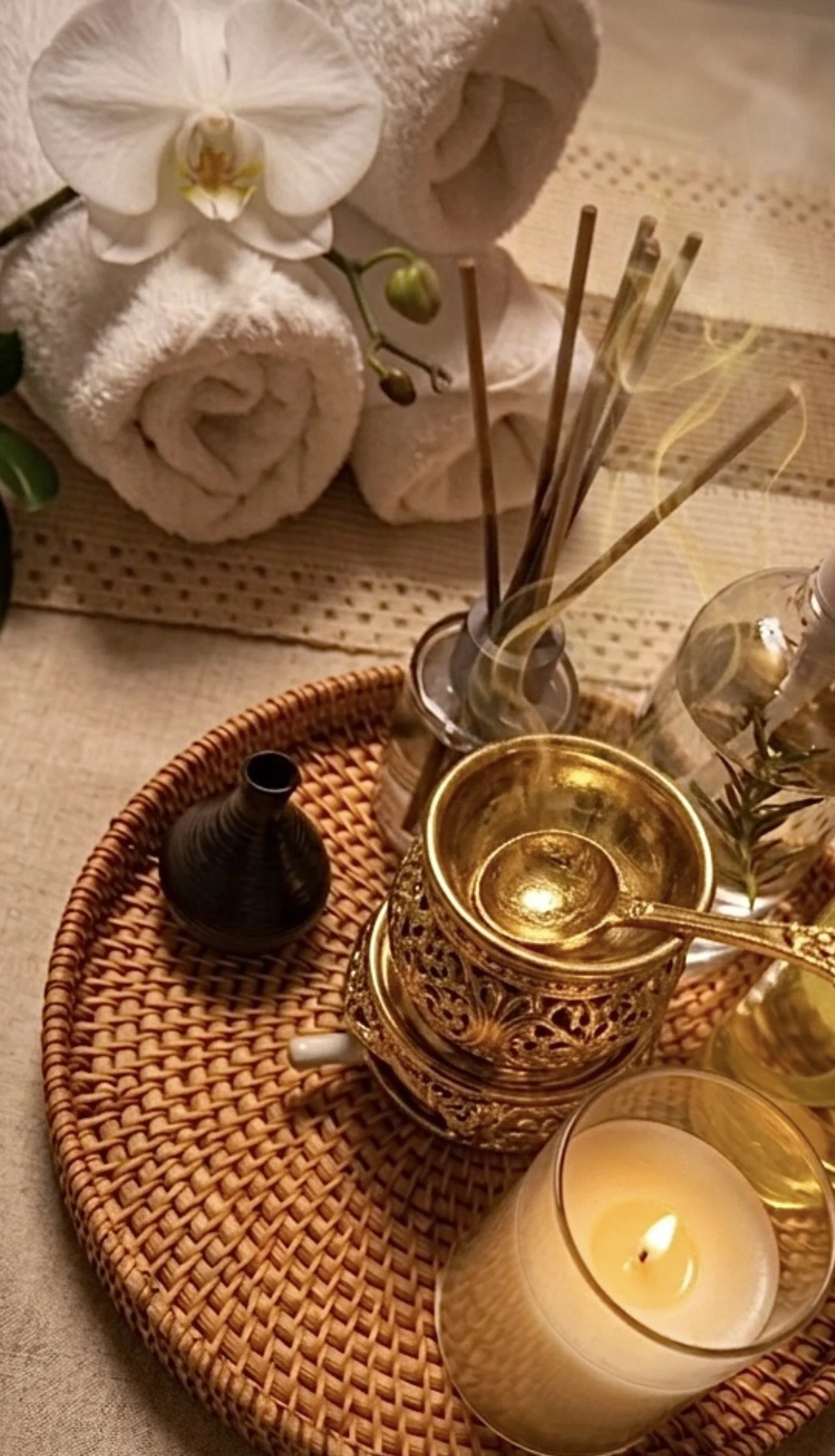 Ein dekoratives Tablett mit einer goldfarbenen Kerze, einem tragbaren Teelicht, einer Glasvase mit kleinen Zweigen, einem dunklen Flaschenvase, einem Wasserkrug mit Strohhalm, einem Glas Wasser und einem Handtuch mit Orchidee, alles auf einem Tisch