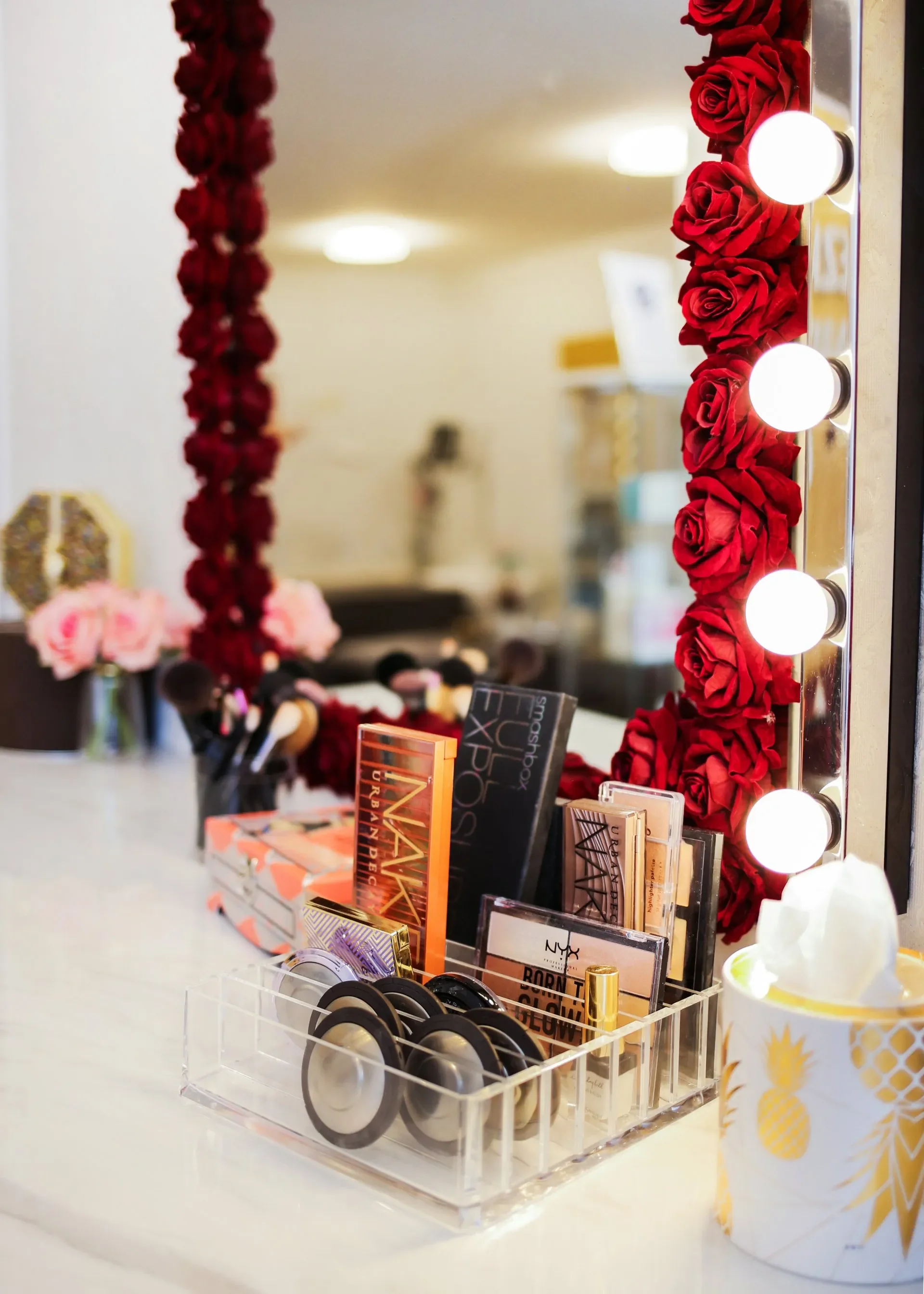 Ein Make-up-Tablett mit verschiedenen Kosmetikprodukten, darunter Lidschatten, Lipgloss und Puder, sowie Makeup-Pinsel. Im Hintergrund sind rote Rosen als Dekoration und ein Spiegel mit Lichterkette.