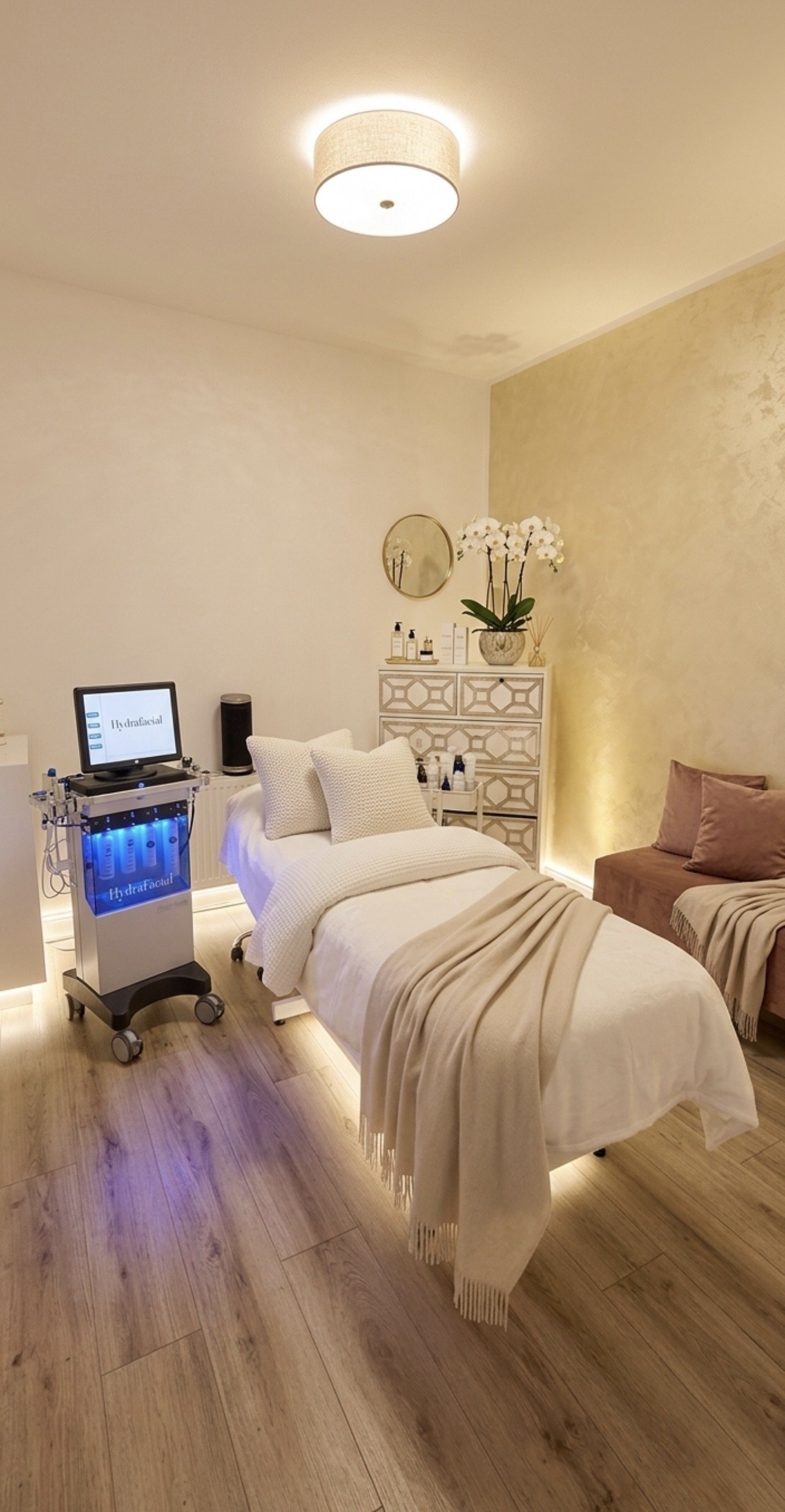 Ein Behandlungsraum mit einem Behandlungsbett, einem kleinen Tisch, einem Kristall-Lichttherapiesystem, einer Kommode mit Pflanzen und dekorativen Gegenständen, einer braunen Couch, hölzernen Boden und weichen Beleuchtung.