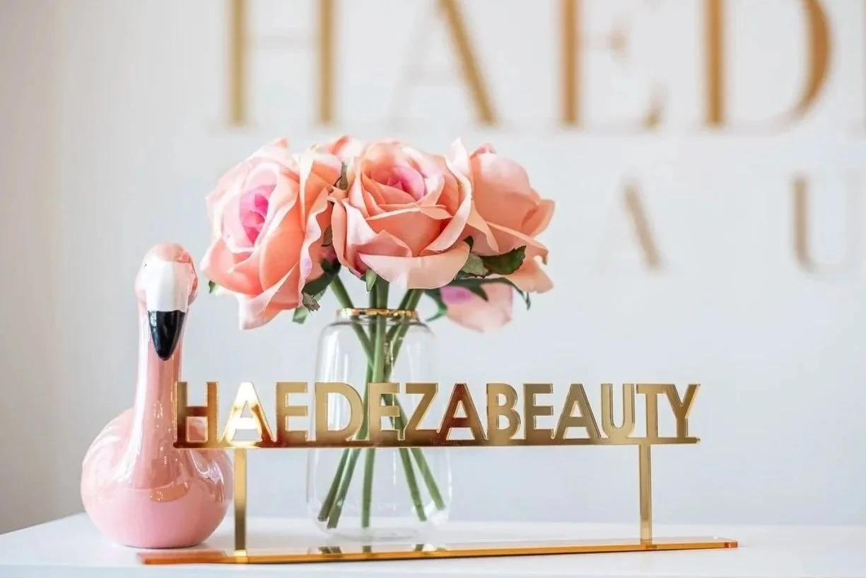 Eine Vase mit pinkfarbenen Rosen, ein rosa Flamingo-Deko und ein goldener Schriftzug mit der Aufschrift "HAEDEZABEAUTY" auf einem weißen Tisch.
