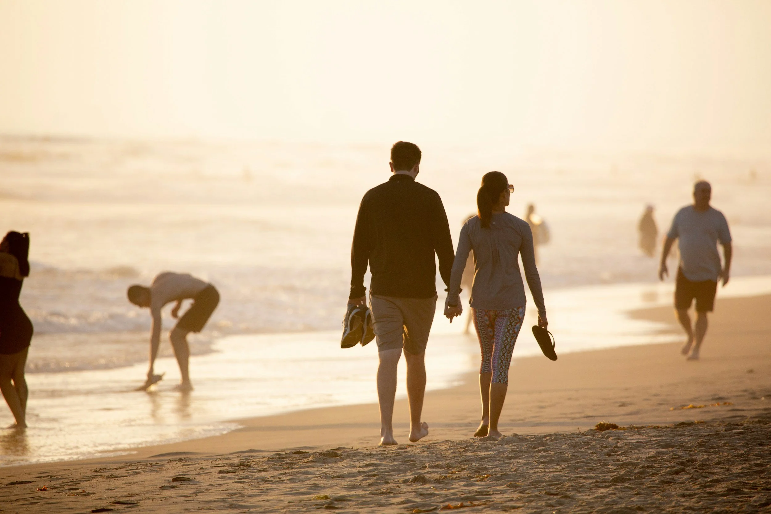Couple walking on the beach during sunset, with other people in the background, some bending down and others strolling along the shoreline.