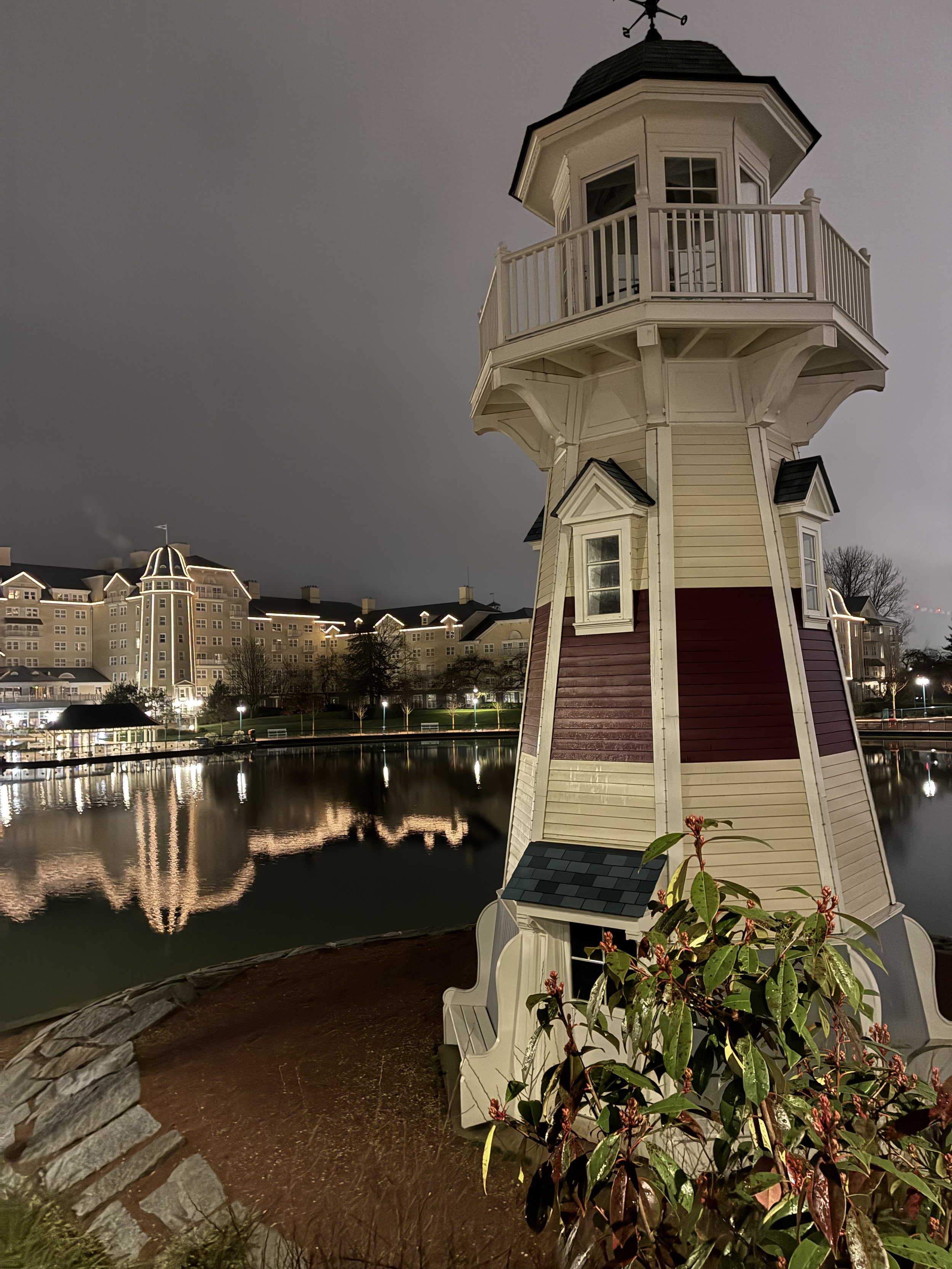 Nighttime view of a whimsical lighthouse beside a body of water with a cityscape in the background. The lighthouse has a red and white exterior, a small covered porch, and a balcony at the top. The water reflects the lights of the buildings and the lighthouse.