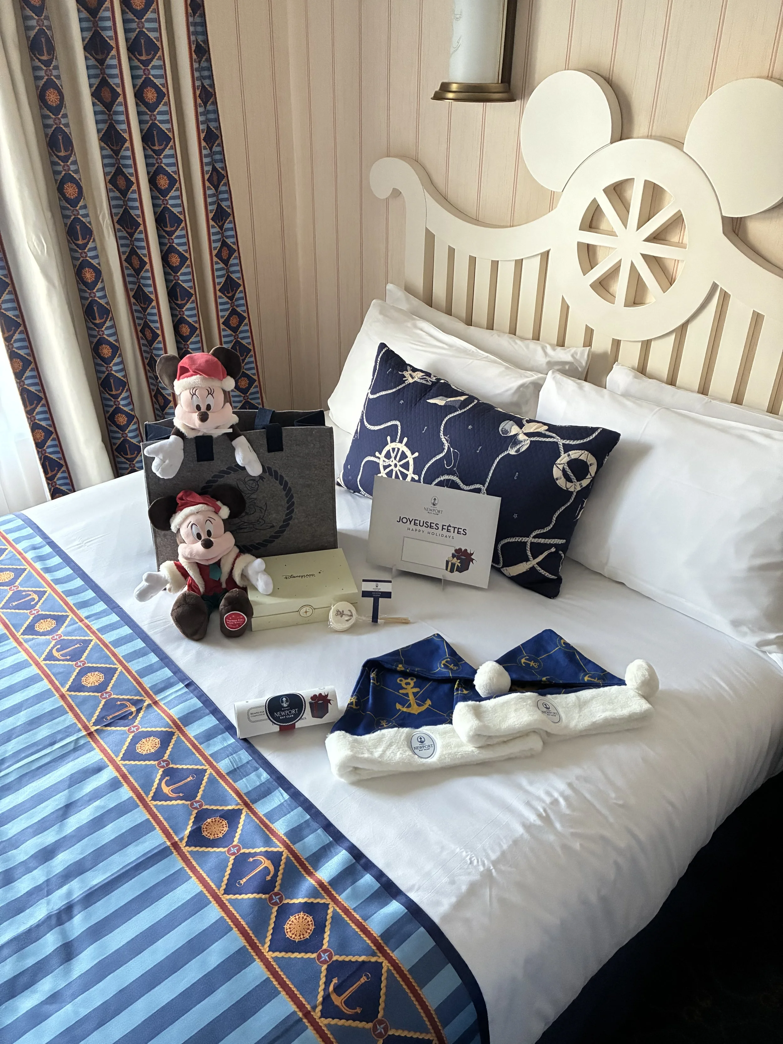 A hotel bed decorated with Minnie Mouse plush toys wearing red Santa hats, a navy blue pillow with nautical designs, and various wrapped gifts and items with nautical and holiday themes. The bed has a white headboard with a circular cutout design, and the room features nautical-themed curtains and wall decor.