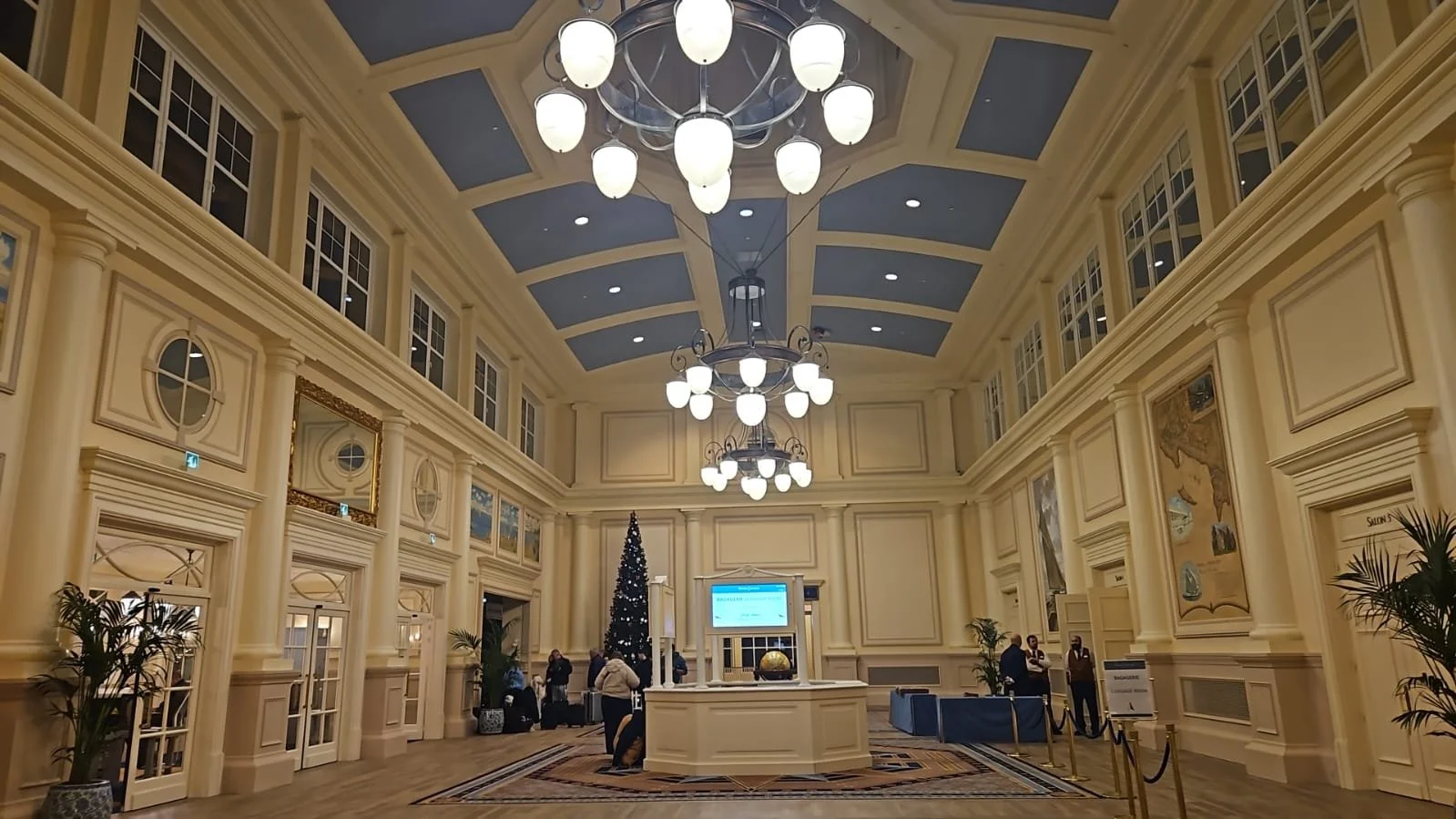 Elegant hotel lobby decorated for Christmas with a tall Christmas tree, chandeliers, cream-colored walls, large windows, and informational displays.