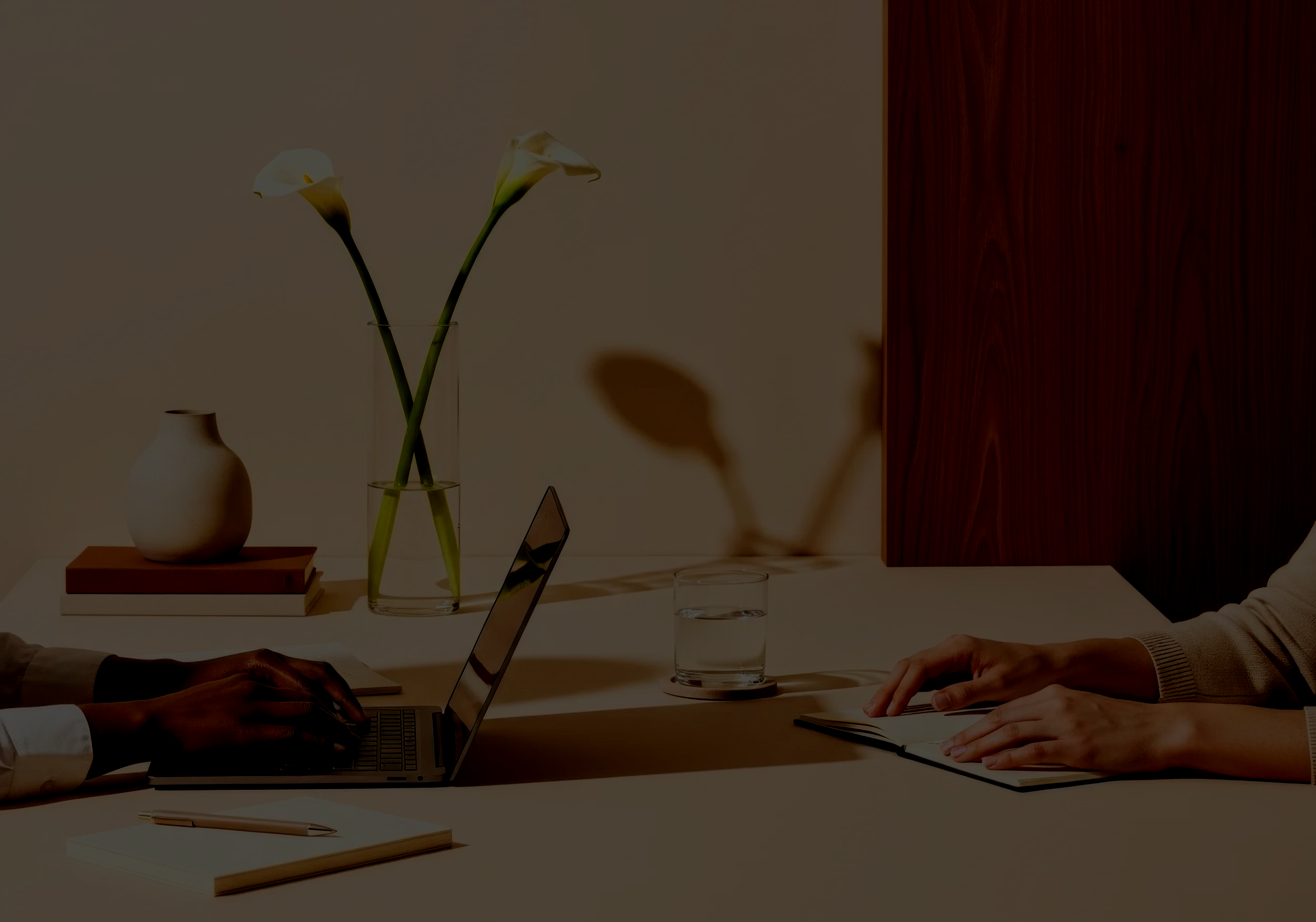 Close-up of two people working at a meeting table, one using a laptop and the other writing in a notebook, with a background featuring a vase with calla lilies and a glass of water.