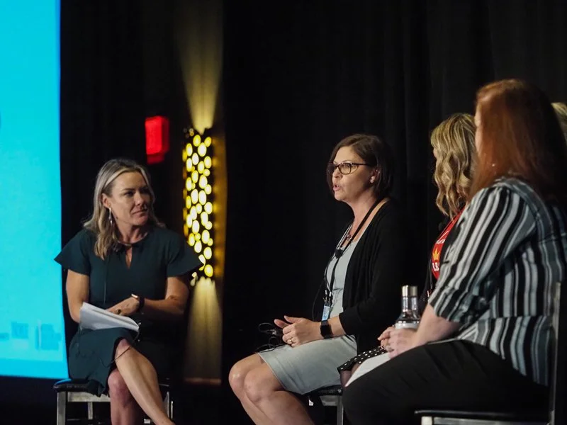 Four women sitting on stage in a panel discussion, with one woman speaking and the others listening, in a dimly lit auditorium.