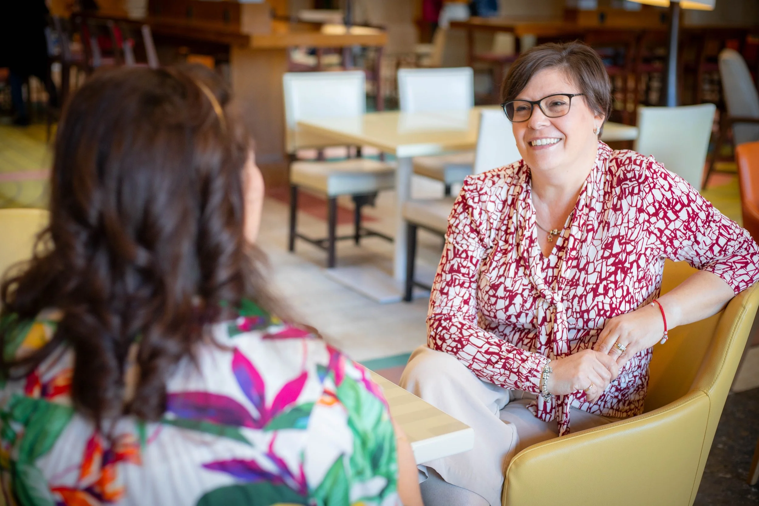 A woman with short brown hair, glasses, and a patterned red and white blouse smiling and talking to another person with long brown hair wearing a colorful patterned top, seated at a table in a restaurant or cafe with wooden furniture.