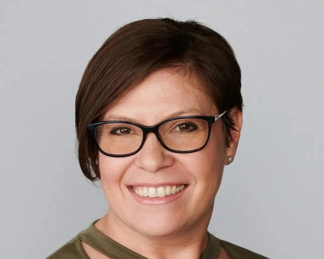 Headshot of a smiling woman with short brown hair, glasses, and earrings, wearing a green top with a round neckline against a gray background.