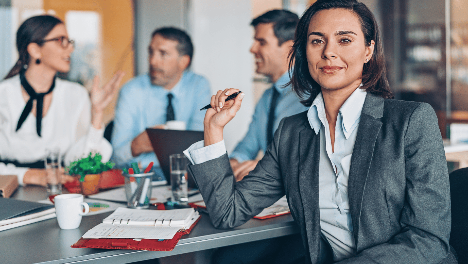 A professional woman in a gray blazer and white shirt sitting at a cluttered table holding a pen, smiling slightly, with colleagues having a discussion in the background.