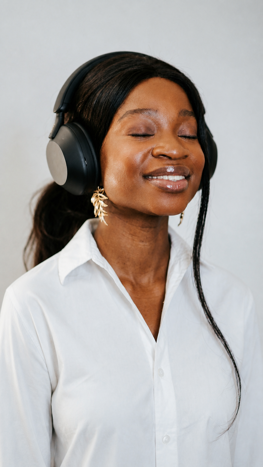 A woman with closed eyes and a gentle smile, wearing large black headphones and a white button-down shirt, standing against a plain light gray background.