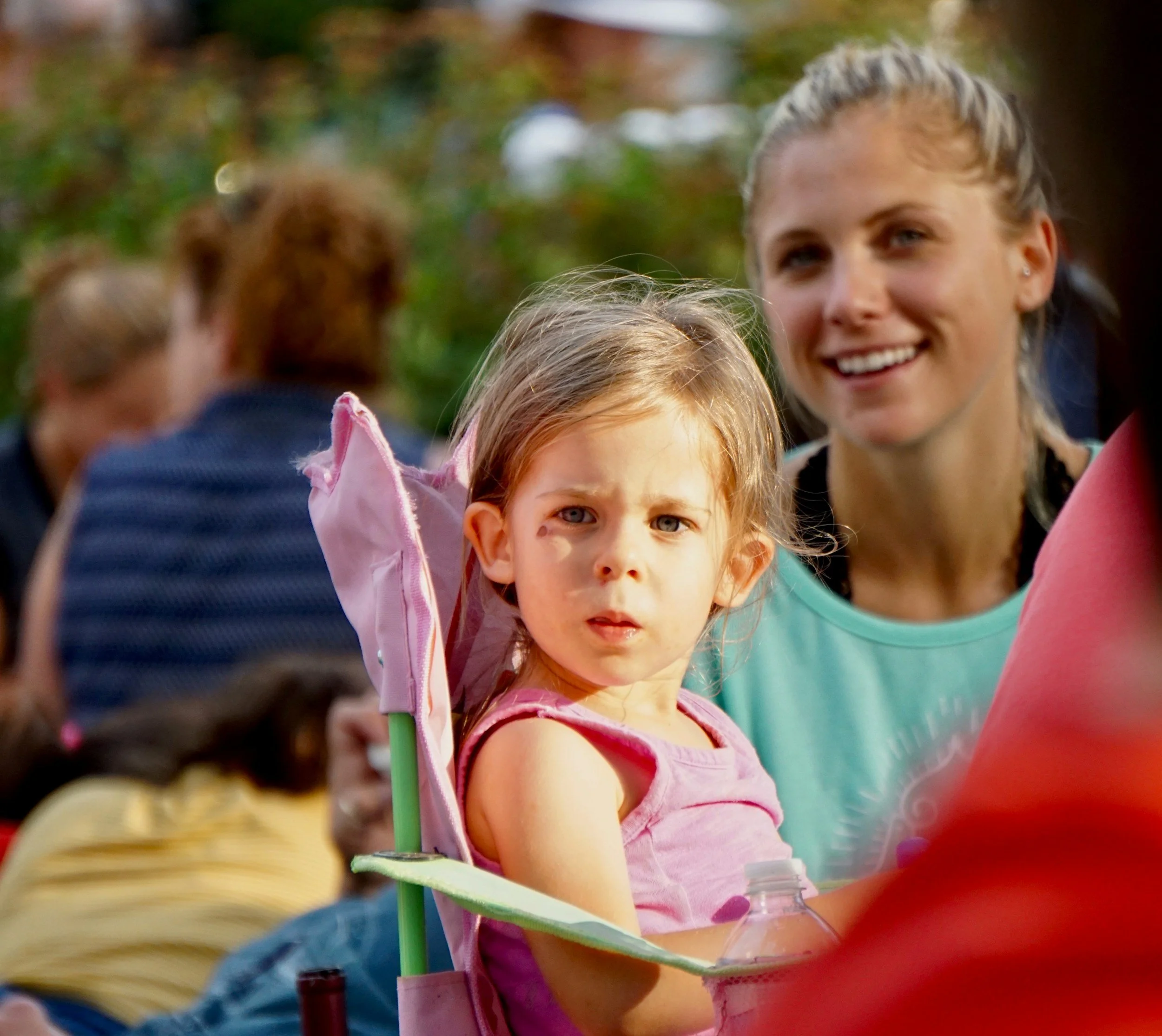 A young girl with blonde hair and blue eyes looking towards the camera, wearing a pink top, outdoor setting with trees in the background. An adult woman with blonde hair is smiling in the background.