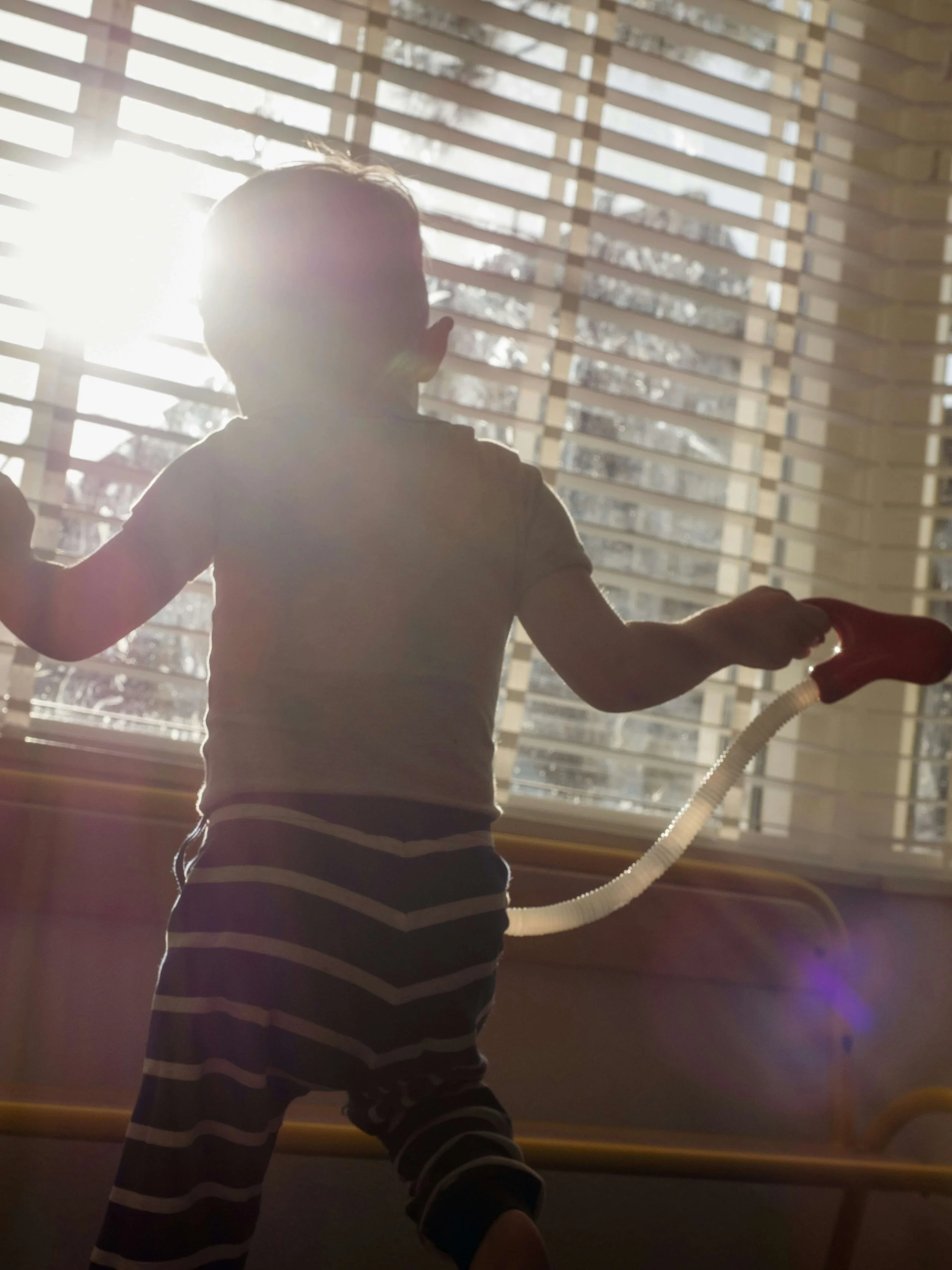 A child wearing a grey and white striped outfit standing in front of a window with horizontal blinds. The sunlight is shining through the window.