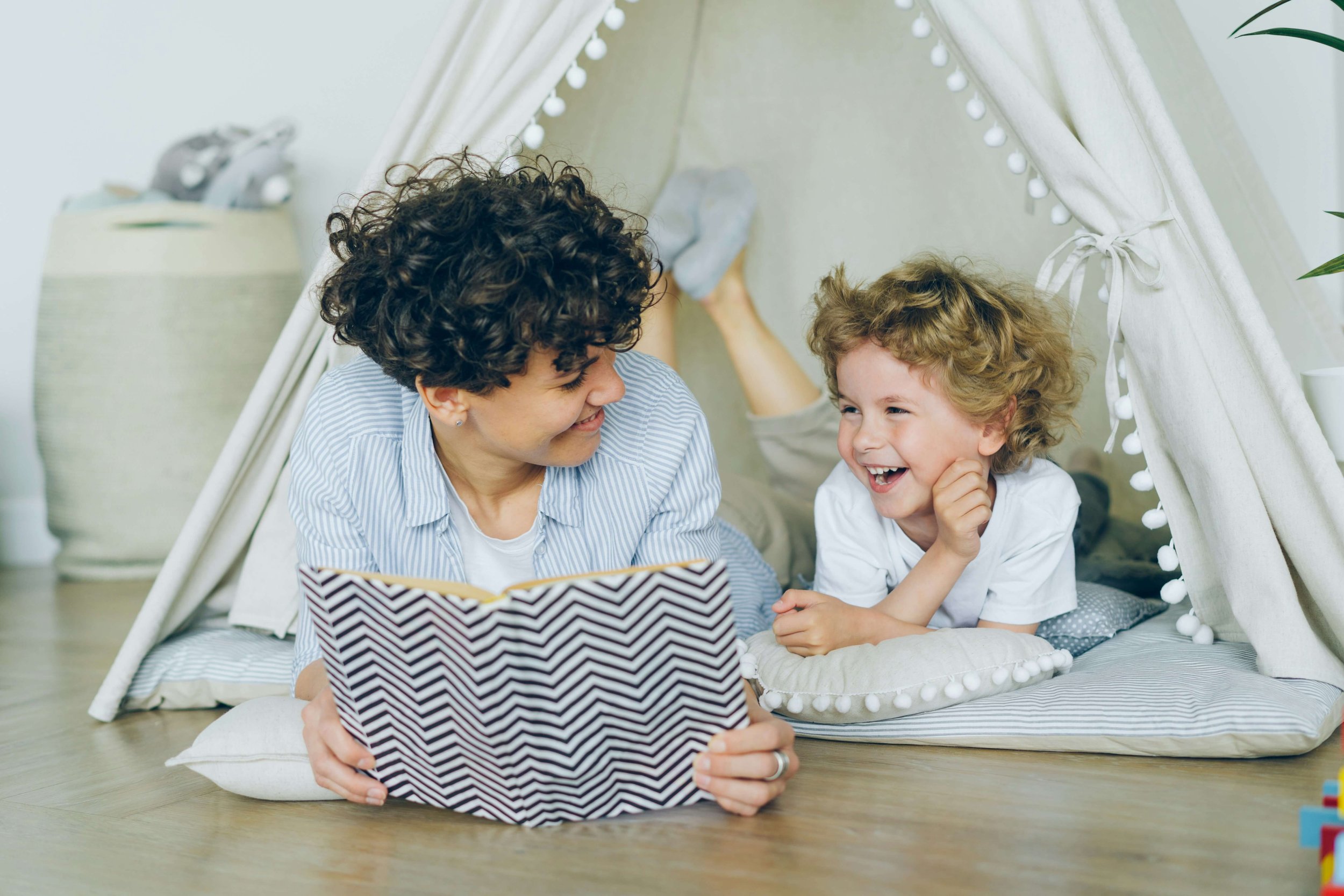 A woman and child lay reading a book on the floor inside a fabric tent, looking at each other and smiling