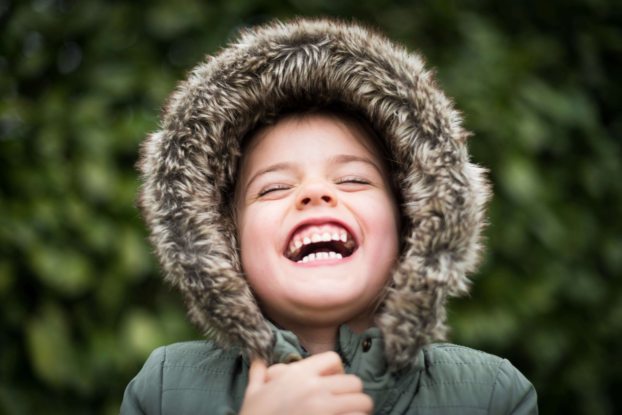 A young child with a broad smile wearing a green coat with a furry hood standing outdoors with a blurred green leafy background.