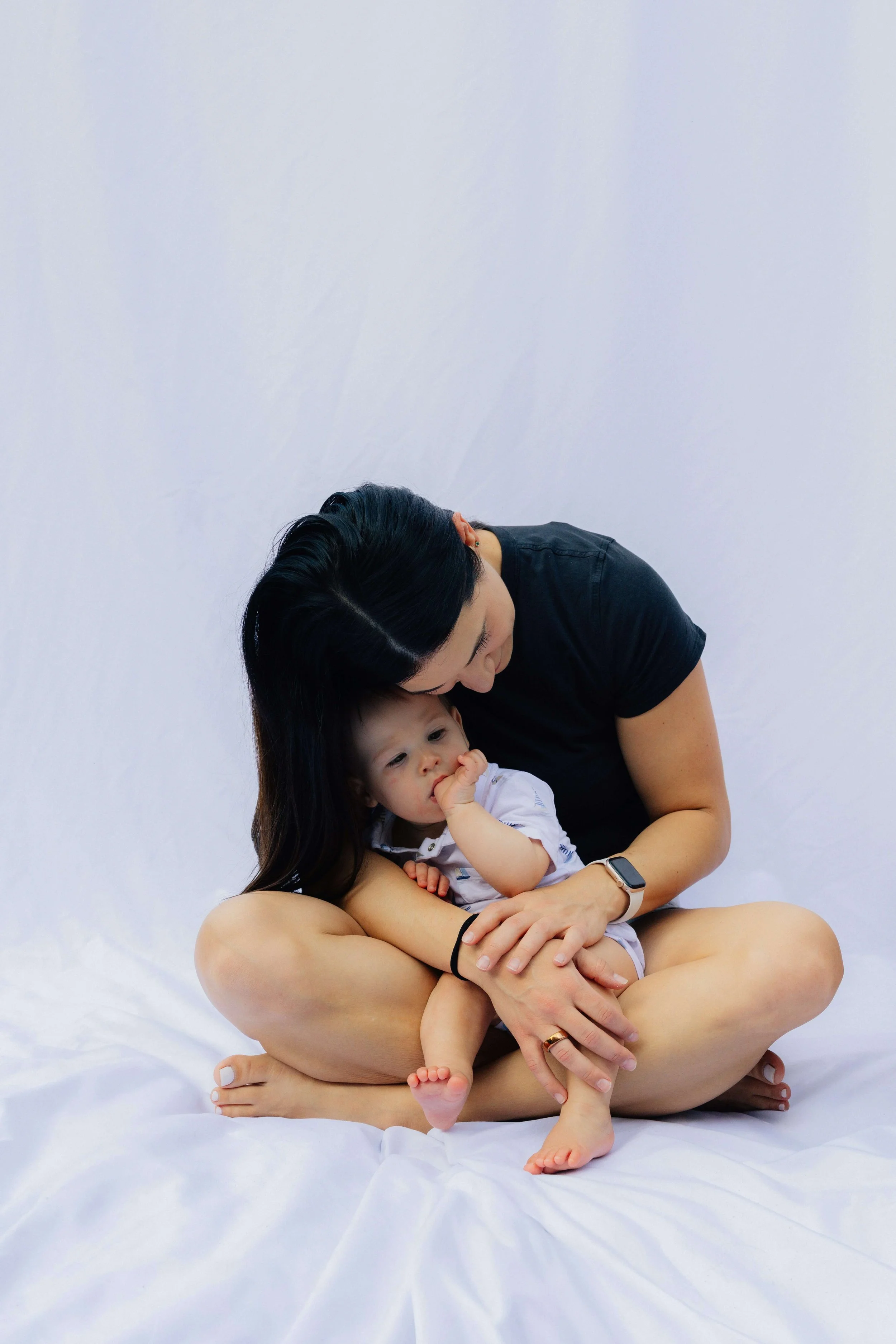 A woman and a baby sitting on the floor against a plain white background, with the woman holding and leaning over the baby.