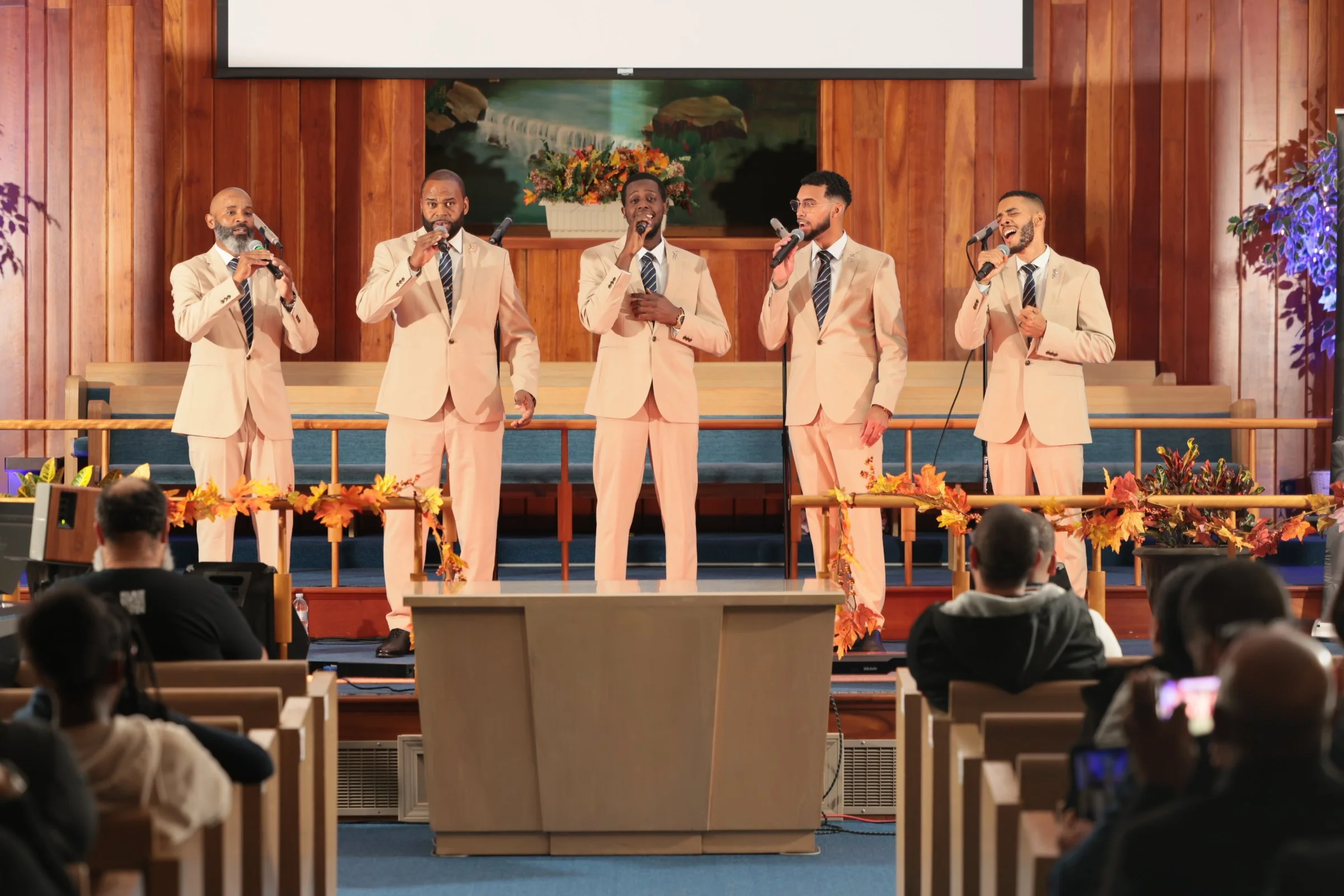 A choir of five men singing on stage in a church, dressed in beige suits with dark striped ties, with a large floral arrangement and a landscape painting in the background, and an audience seated in pews in front.
Heralds of freedom 4th annual concer