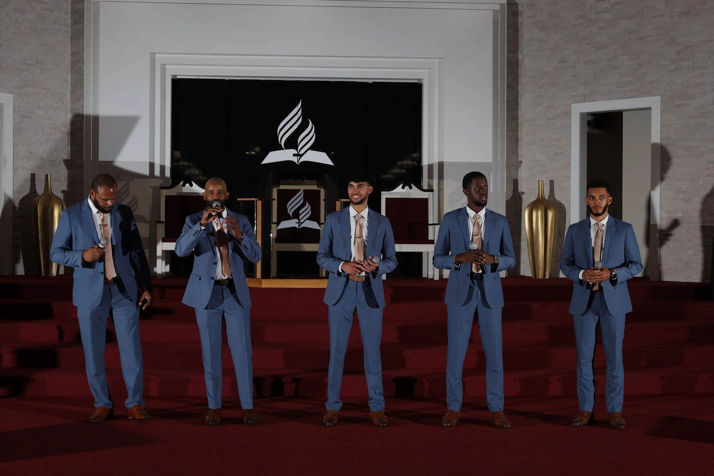 Five men in blue suits and beige ties standing on a red-carpeted stage with microphones, in front of a black background with a white flame logo over an open book, in a tastefully decorated room with large gold vases.
Heralds of freedom 3rd annual con