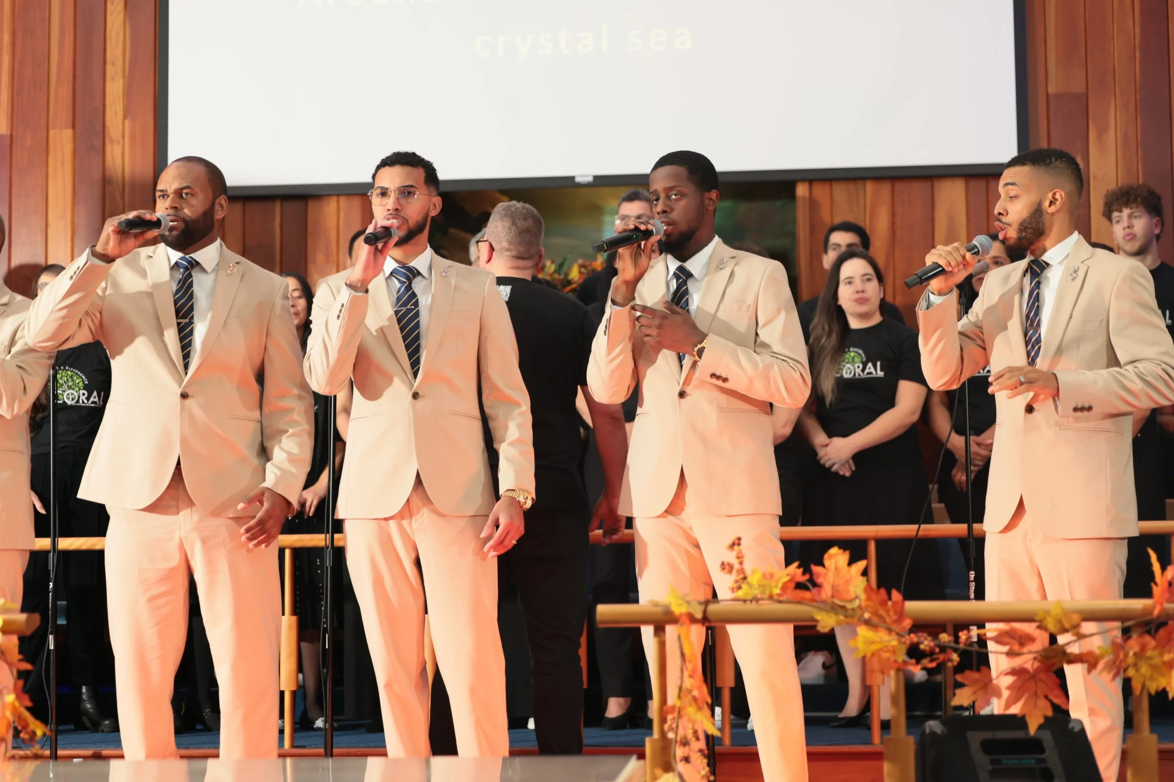 Four men dressed in beige suits with striped ties standing on a stage, holding microphones, in front of a choir, with a large screen behind them displaying the words 'crystal sea.'
Heralds of freedom 4th annual concert.