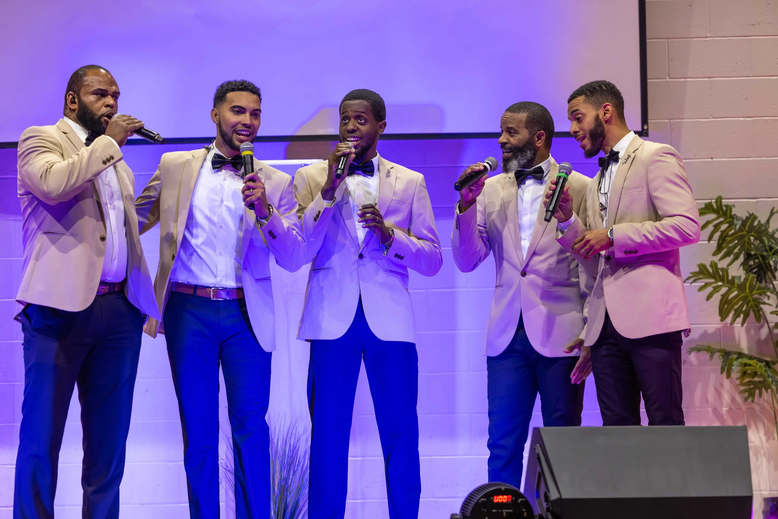 A group of five men dressed in beige tuxedos with black bow ties are singing into microphones on stage, with purple lighting and a white brick wall in the background.
Heralds of freedom 5th annual concert.