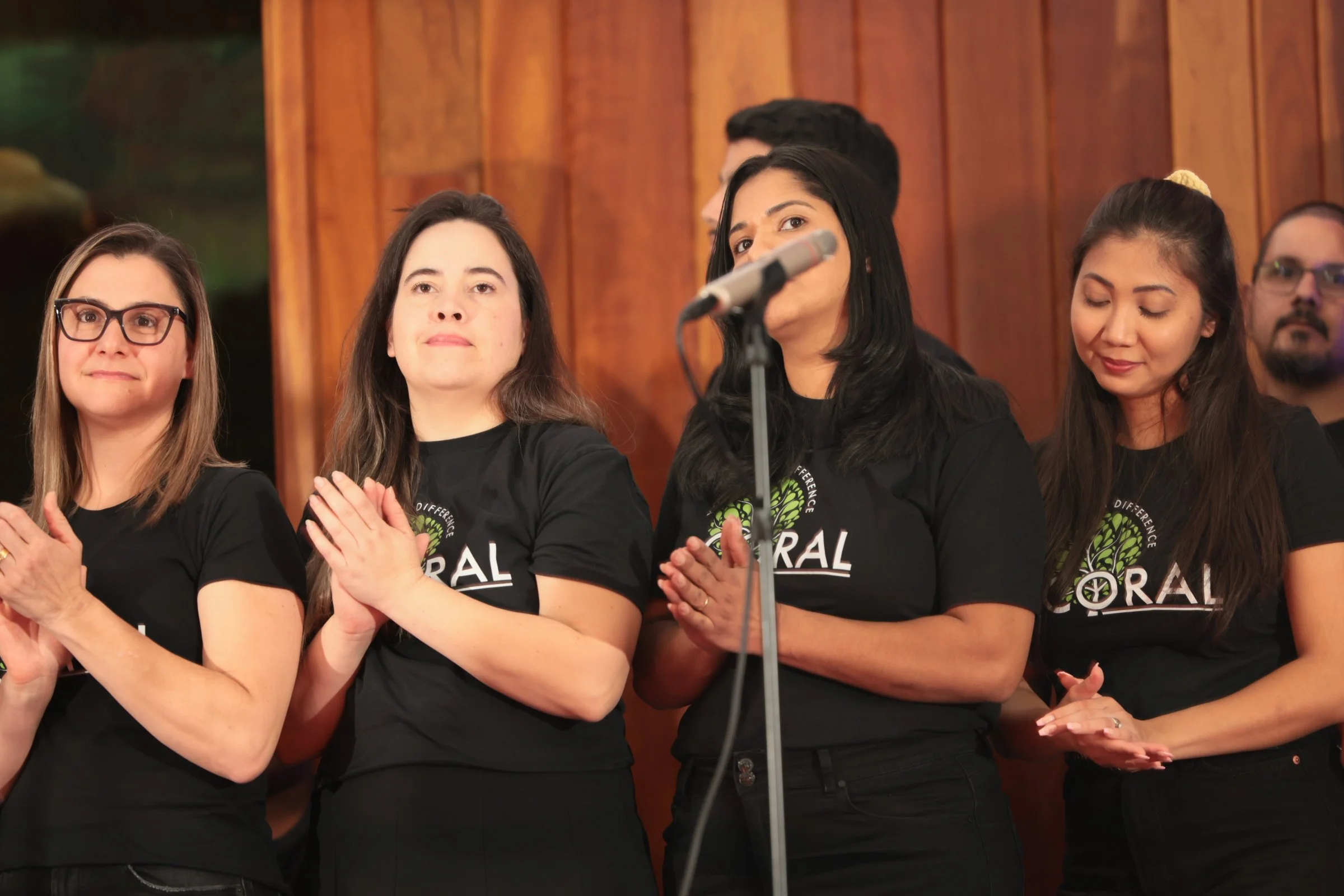 A group of diverse women standing in a row, clapping, with a wooden panel wall in the background. They are wearing black T-shirts with a green and white logo and the words 'Different Single'. They appear to be at an event or presentation, and one wom
