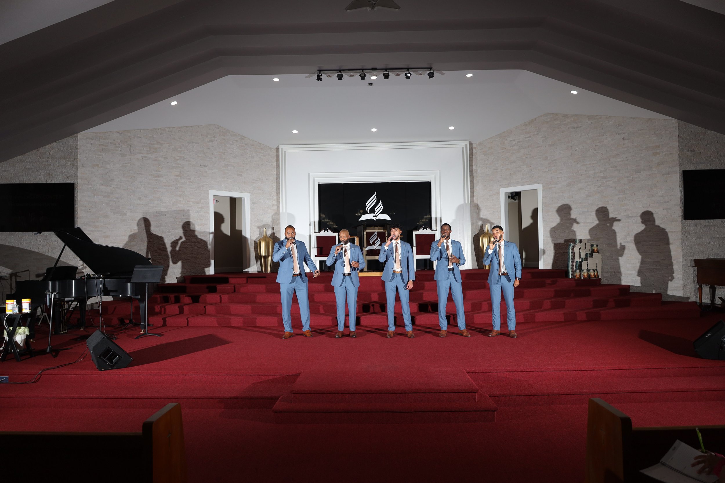 A group of six men dressed in matching blue suits and brown shoes singing on a stage with a red carpet. There are stage lights and a piano on the left side, with a dark background and a logo on the wall behind them.
Heralds of freedom 3rdannual conc