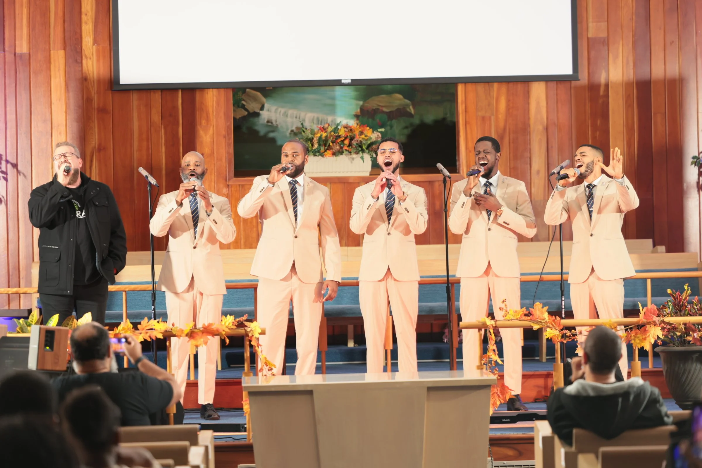 A choir of six men singing on stage with wooden paneling and a mountain landscape painting in the background, decorated with fall foliage flowers.
Heralds of freedom 4th annual concert.