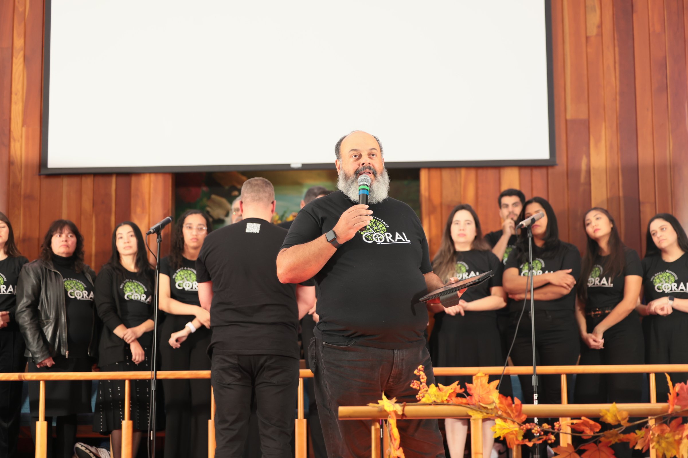 A group of people standing on a stage with wood paneling, led by a man with a beard holding a microphone. The crowd appears to be at an event, with autumn decorations and a large blank screen behind them.
Heralds of freedom 4th annual concert.