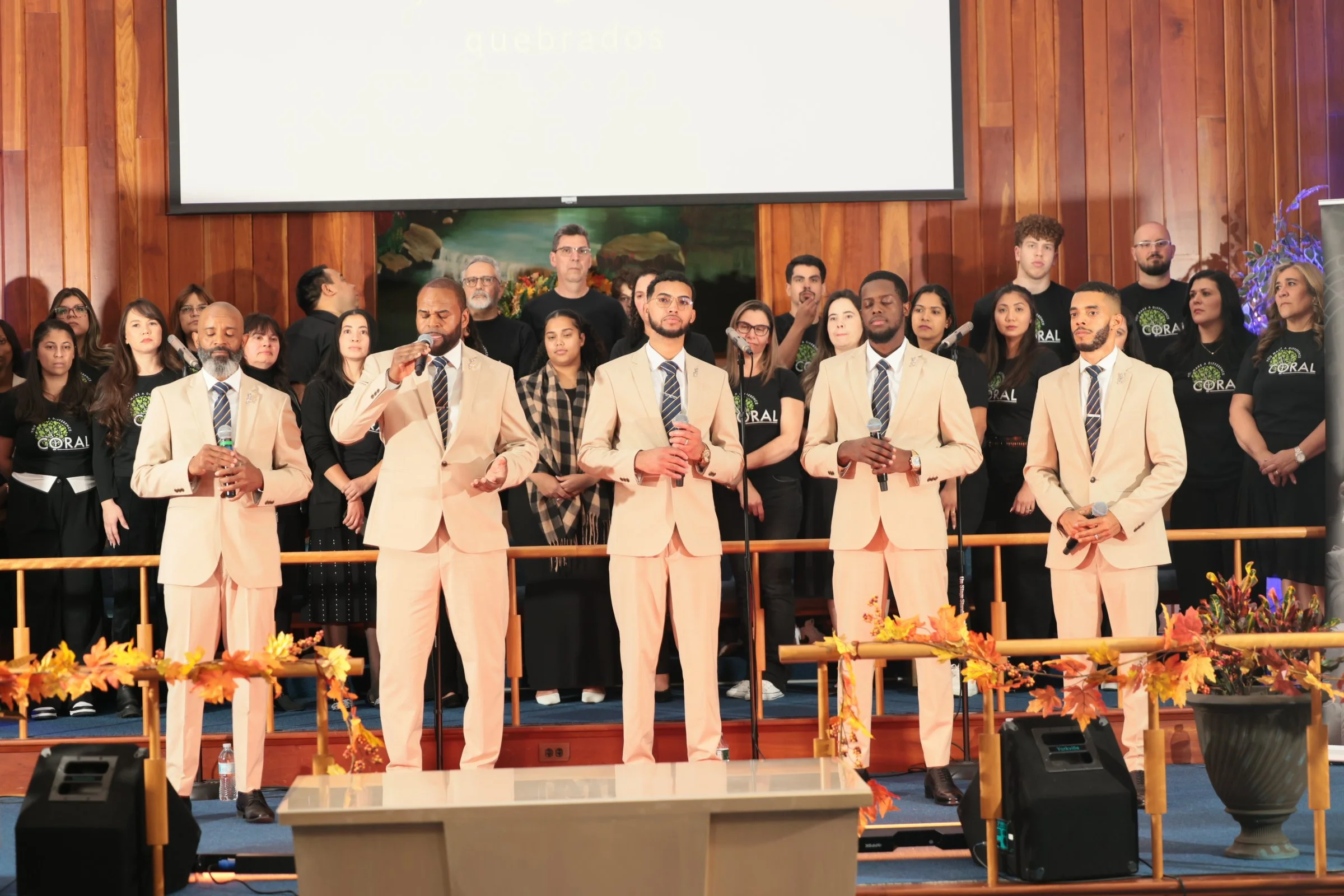 A choir performing on stage at a church or auditorium, with a group of men and women dressed in formal and casual attire, some holding microphones, decorated with autumn-themed flowers and foliage.
Heralds of freedom 4th annual concert with You make 