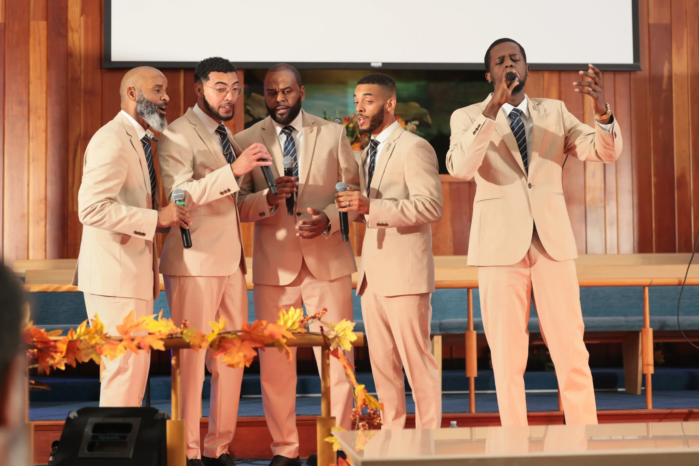 Five men dressed in beige suits with dark ties singing into microphones on stage, with a large white screen and wooden-paneled background behind them, and autumn leaves decoration in the foreground.
Heralds of freedom 4th annual concert.