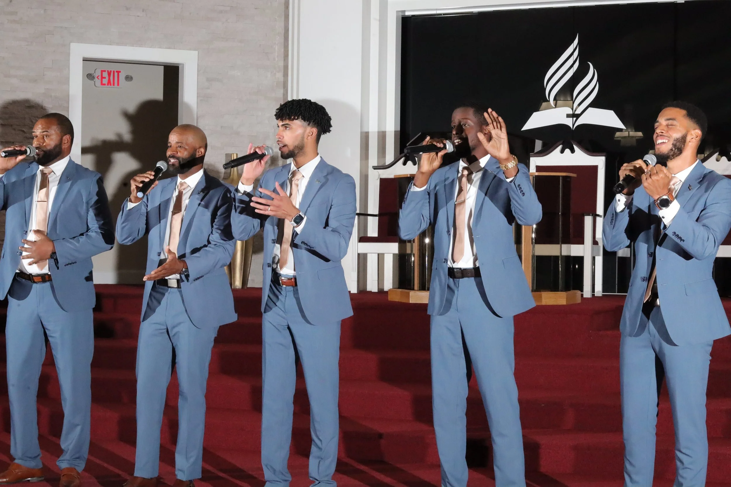 A group of five men dressed in blue suits and ties, holding microphones and singing or speaking on stage in a church or auditorium with a red carpeted stage and a large open Bible and torch logo in the background. Heralds of freedom 3rd annual concer