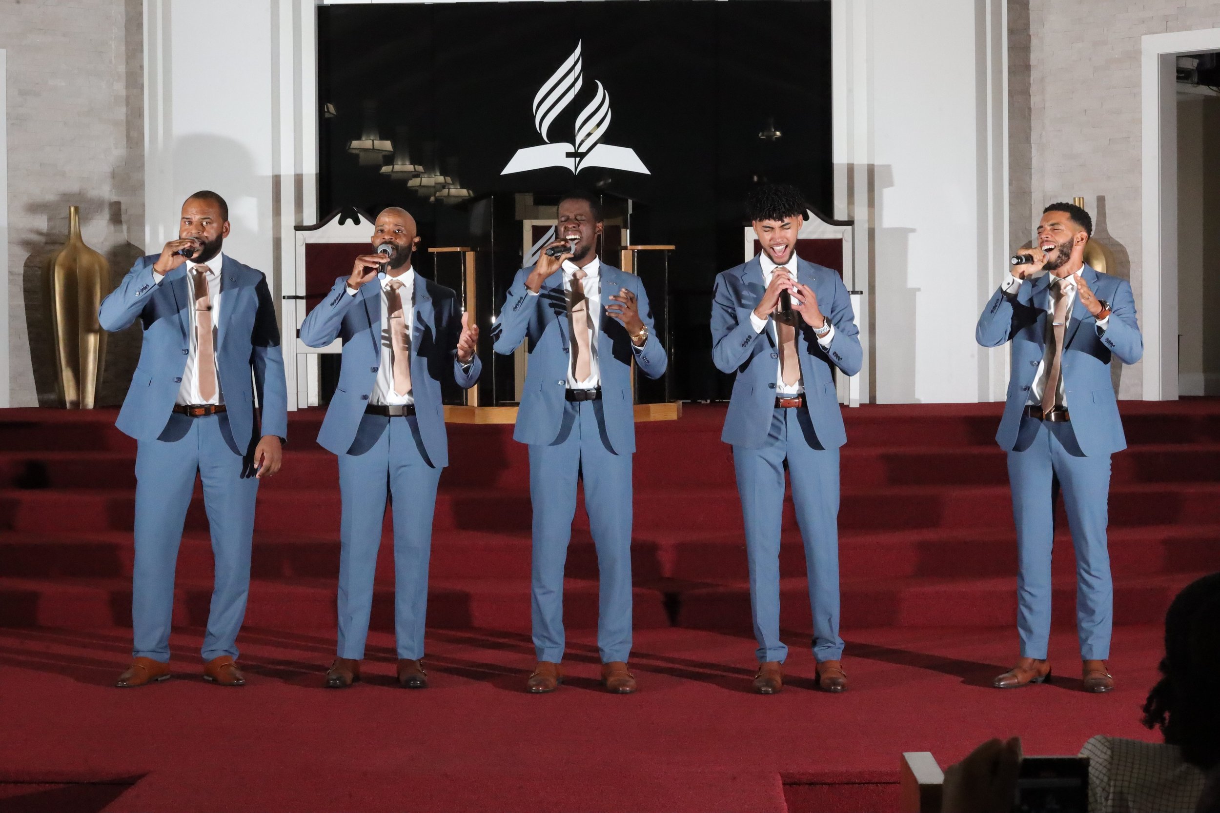 Five men in matching blue suits and tan ties singing into microphones on a stage with a black and white logo of a feather and book behind them. Heralds of freedom 3rd annual concert.