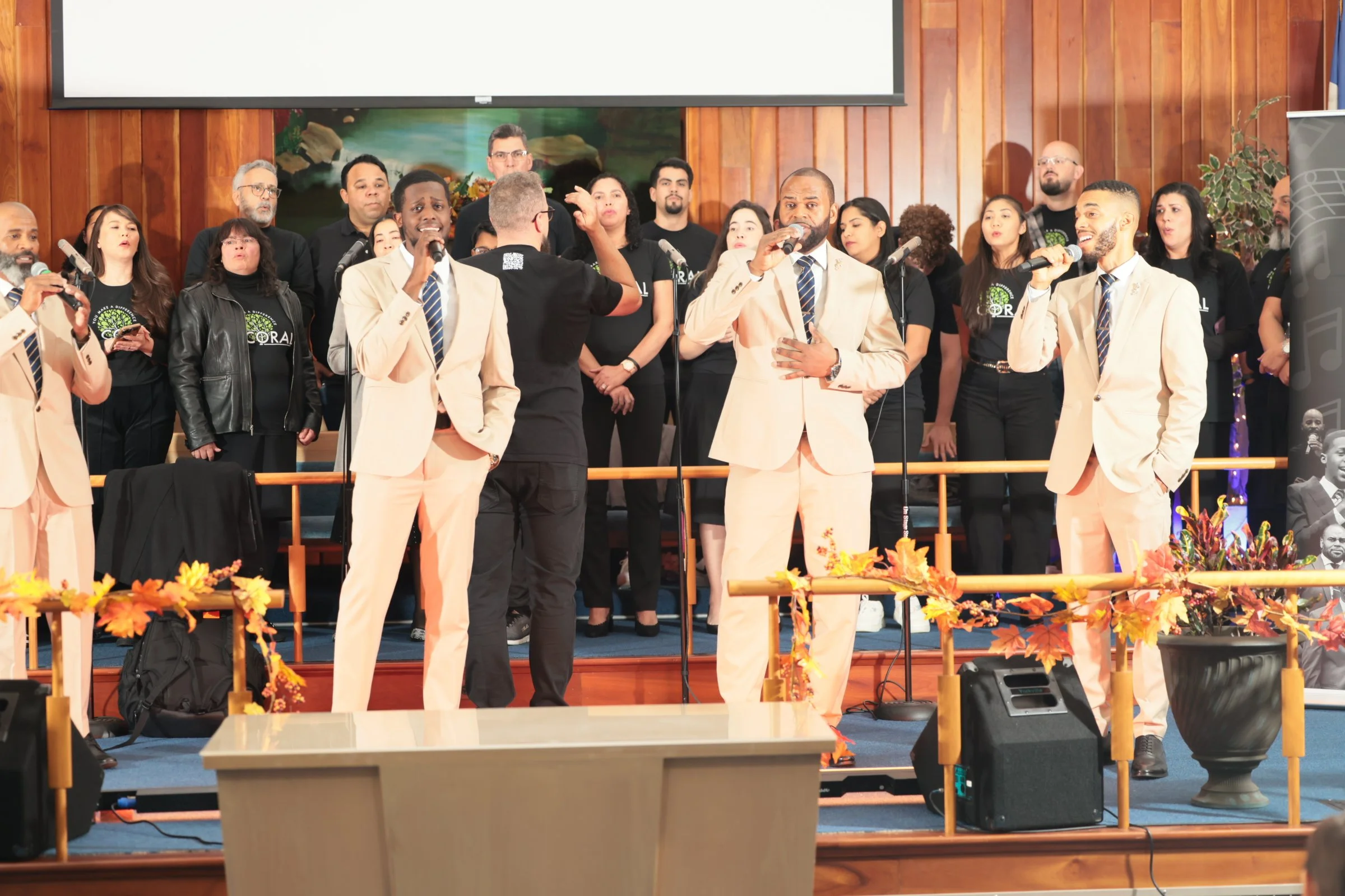 A diverse choir sings on stage during a church service, led by a man in a suit holding a microphone, with wooden paneling and autumn decorations in the background.
Heralds of freedom 4th annual concert with You make a difference coral