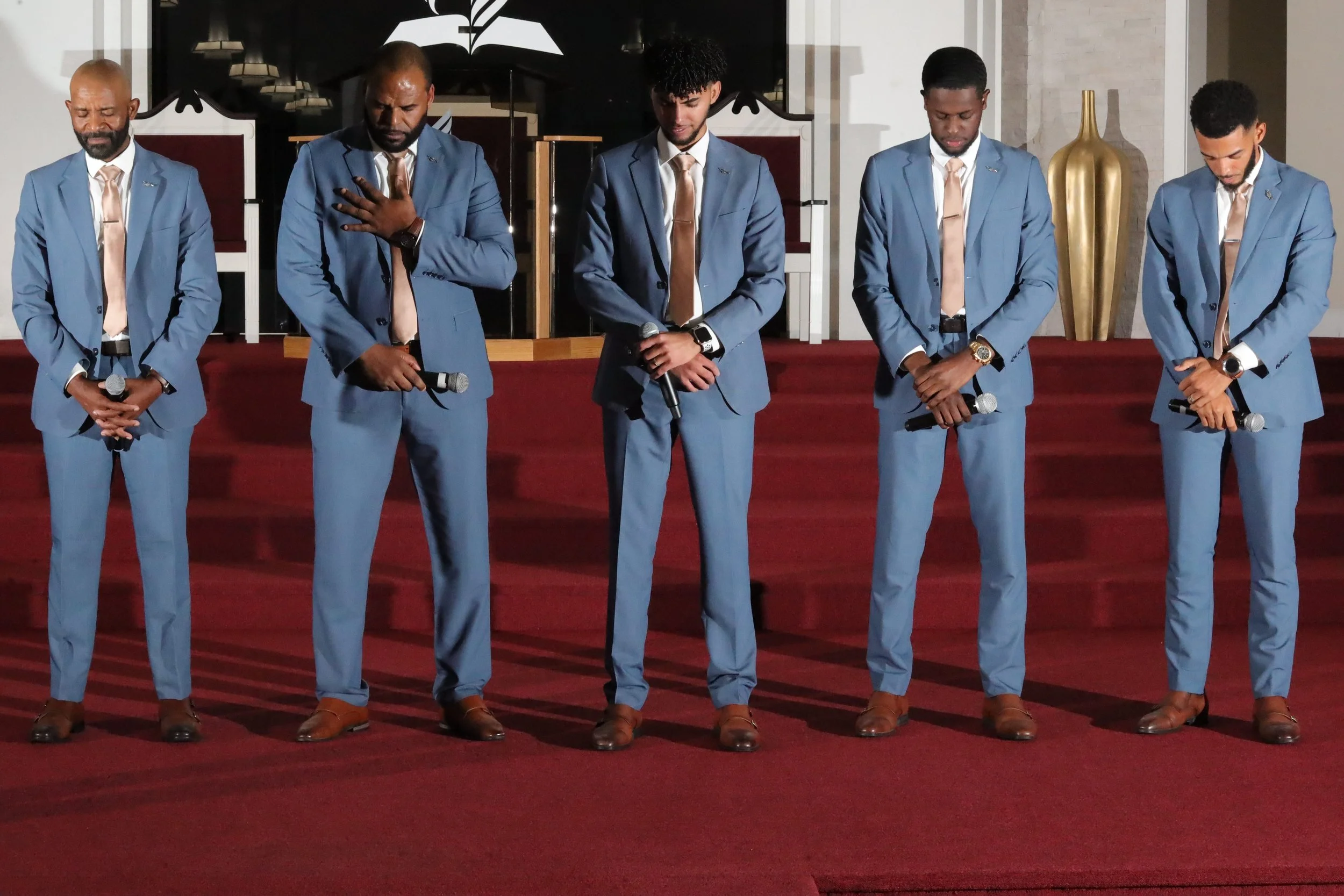 Five men in blue suits with tan ties and brown shoes standing on a stage, holding microphones with heads bowed and eyes closed, in a church or conference hall setting. Heralds of freedom 3rdannual concert.
