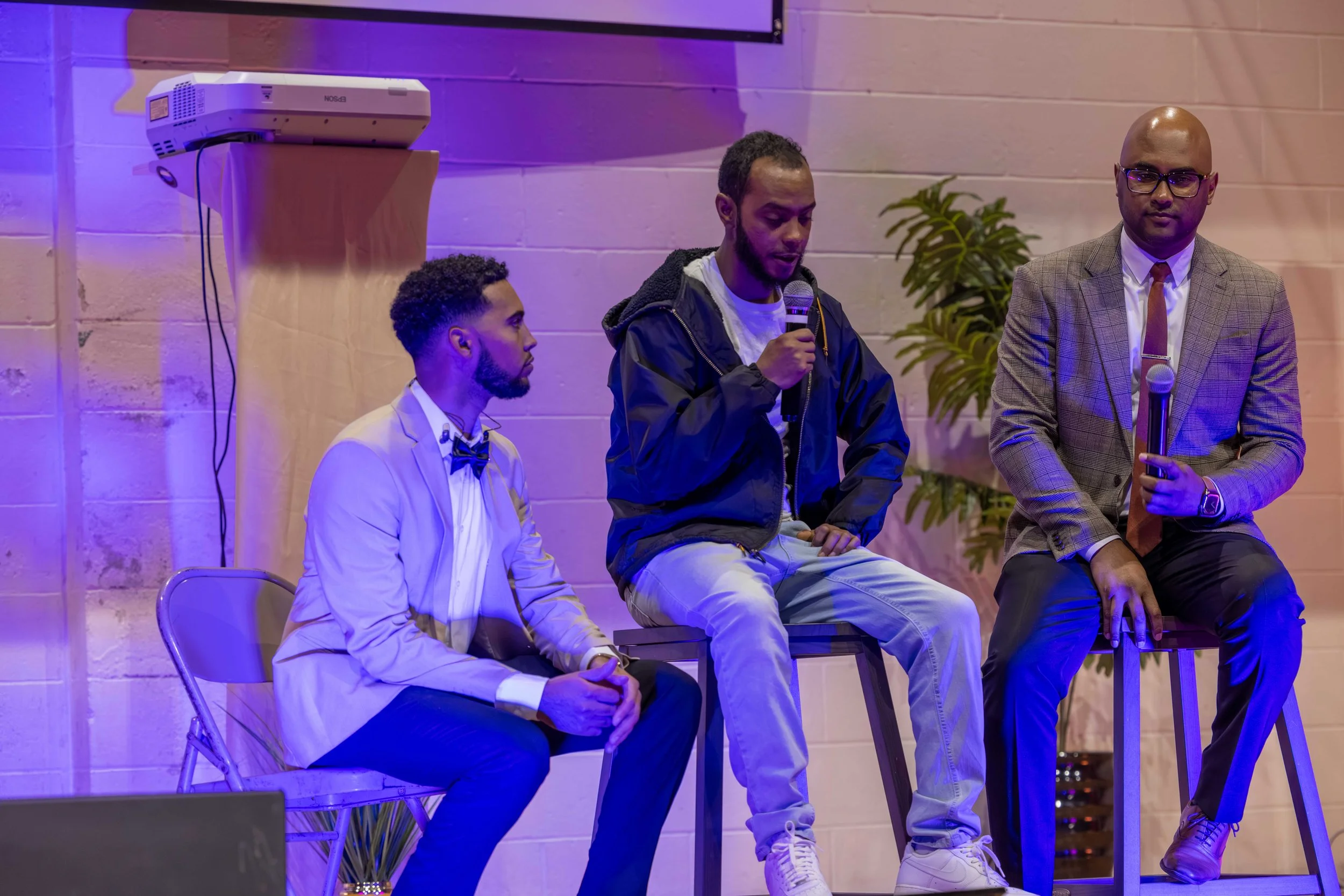 Three men participating in a panel discussion or presentation in an indoor venue with purple lighting. The man in the middle is speaking into a microphone, the man on the right is holding a microphone and dressed in a plaid blazer, and the man on the