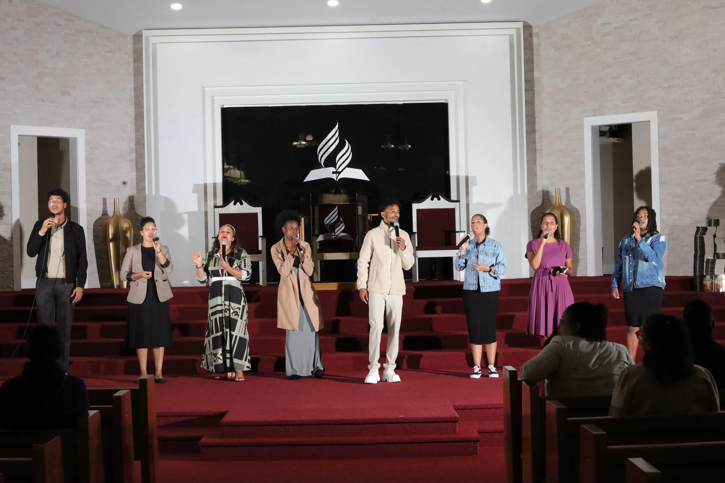 Group of eight people standing on a church stage, singing into microphones, with a black and white logo of a burning flame above an open book in the background. Heralds of freedom 3rd annual concert.