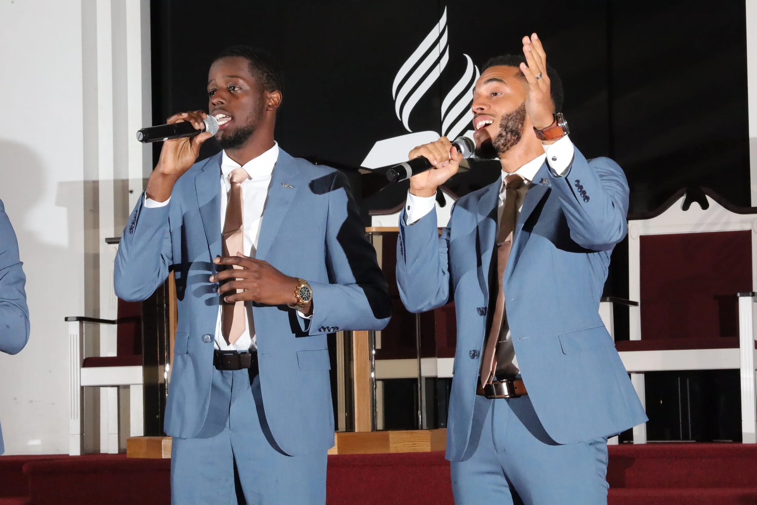 Two men in blue suits singing into microphones on stage, with a black background and church seating. Heralds of freedom 3rd annual concert.