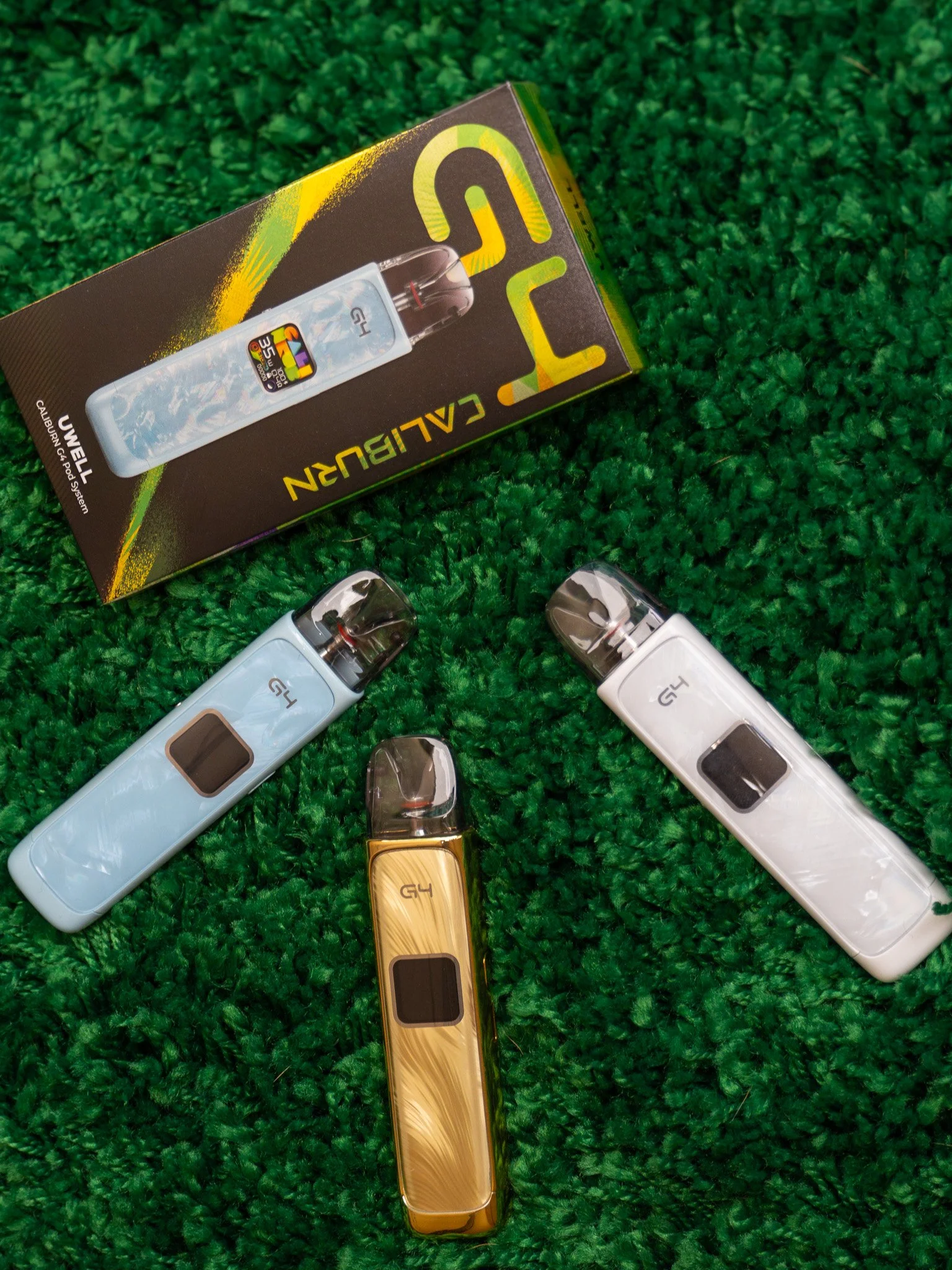 Collection of vape pens and a box of SMYUCALIBURN gadget on green textured surface.