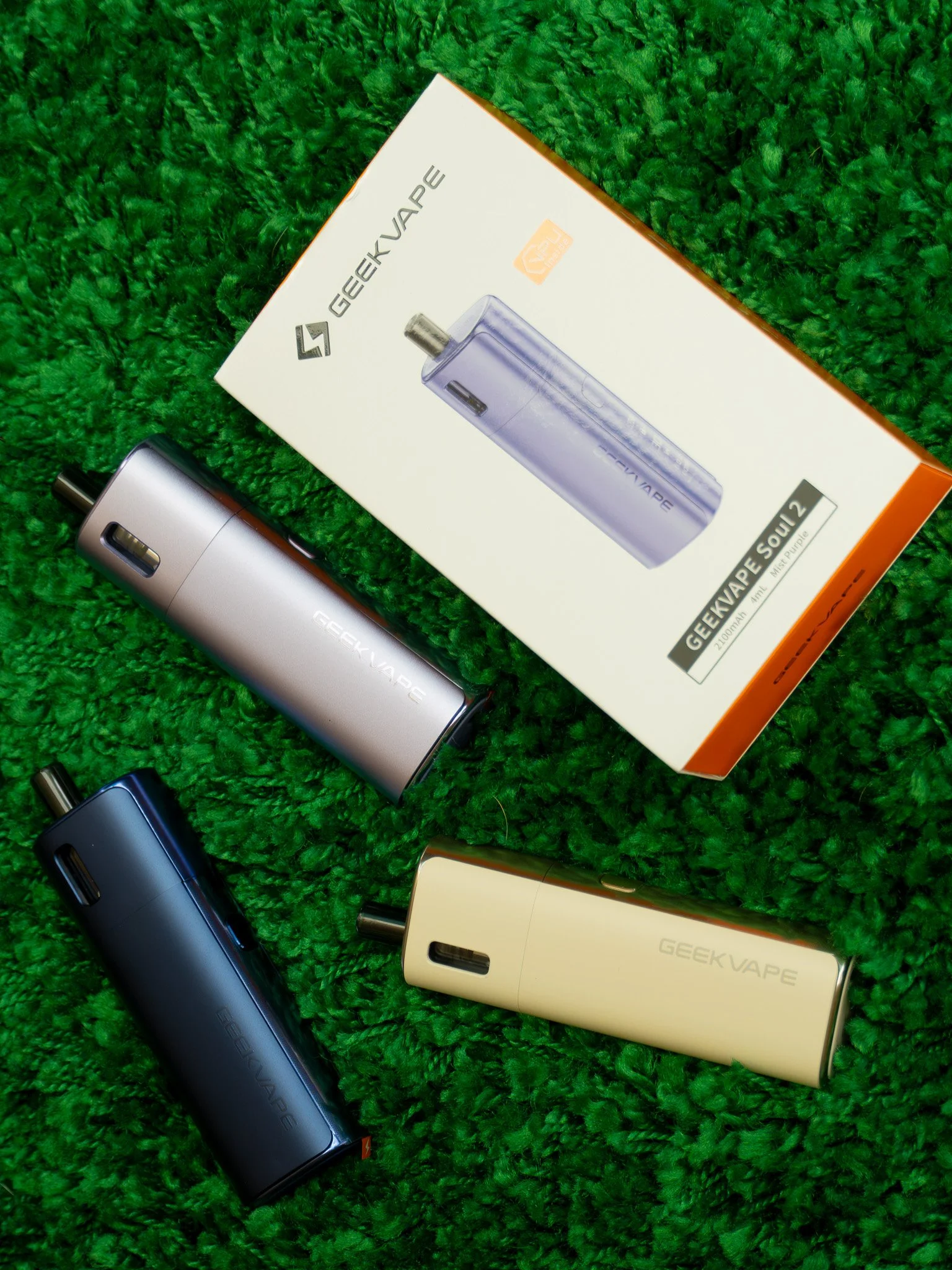 Multiple GeekVape GEEKVAPE electronic vaping devices in black, blue, and gold colors arranged on a green fuzzy surface, alongside boxes with product branding.