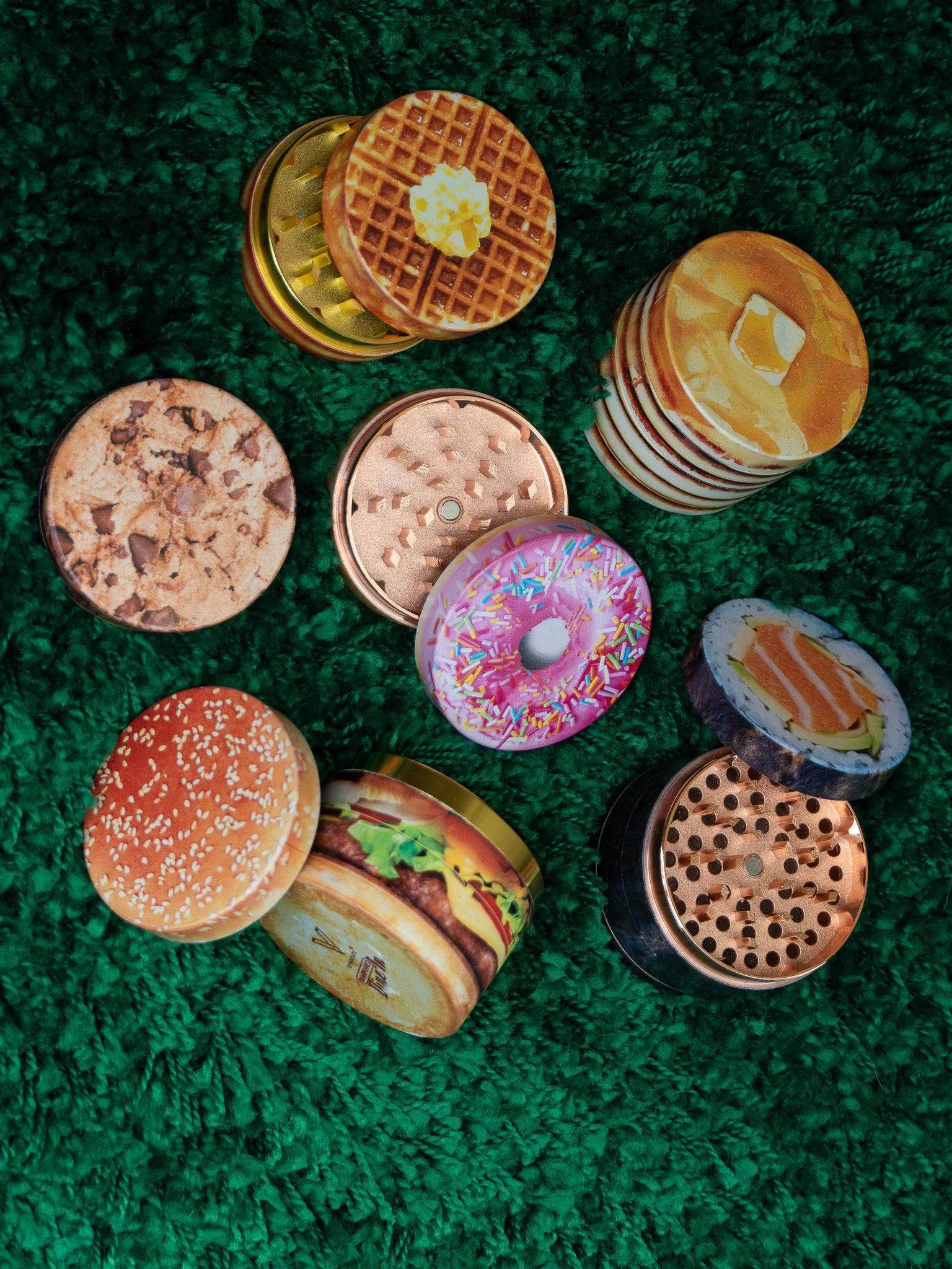 Close-up of a colorful herb grinder with a waffle pattern on top and some popcorn in the center, placed on a textured green surface. There are also other grinders and a sushi roll with salmon and avocado visible in the background.