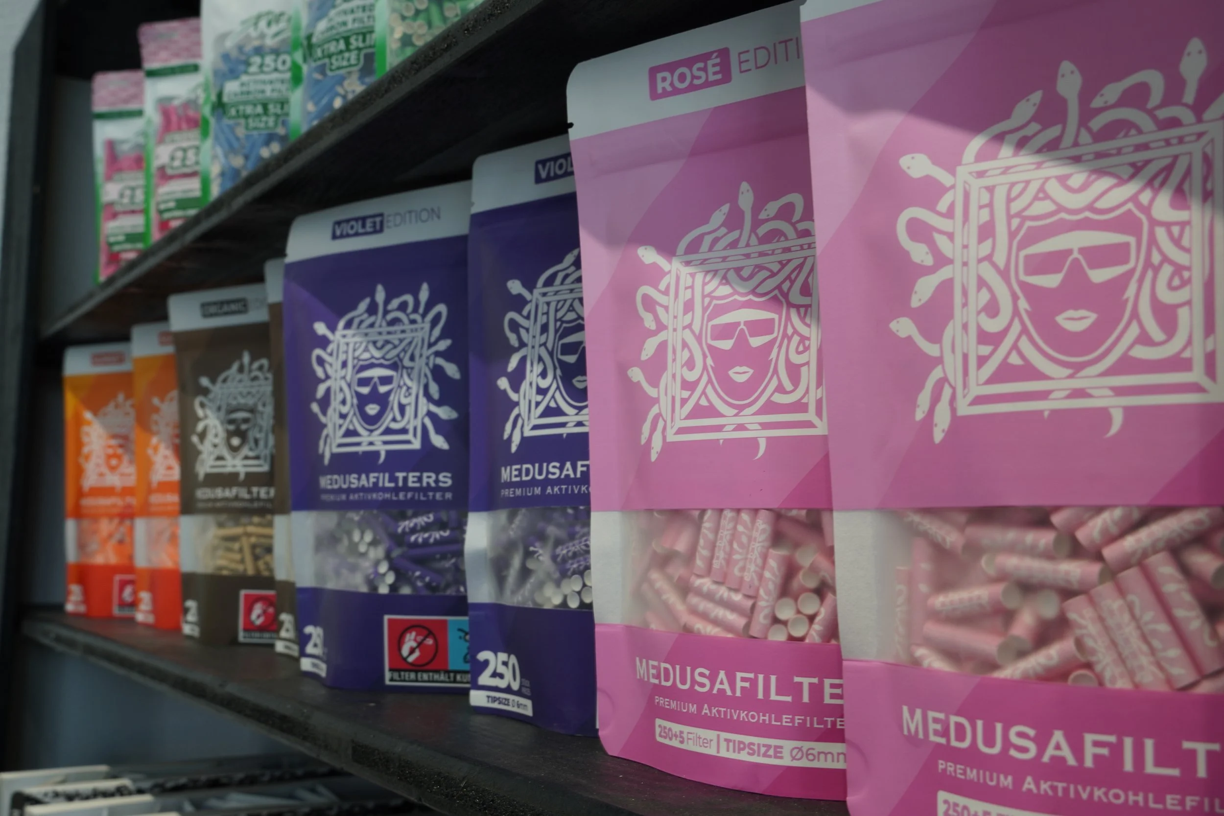 Shelf with packages of Medusa filters, with pink, purple, orange, and blue packaging featuring a stylized woman's face with snakes for hair and goggles.