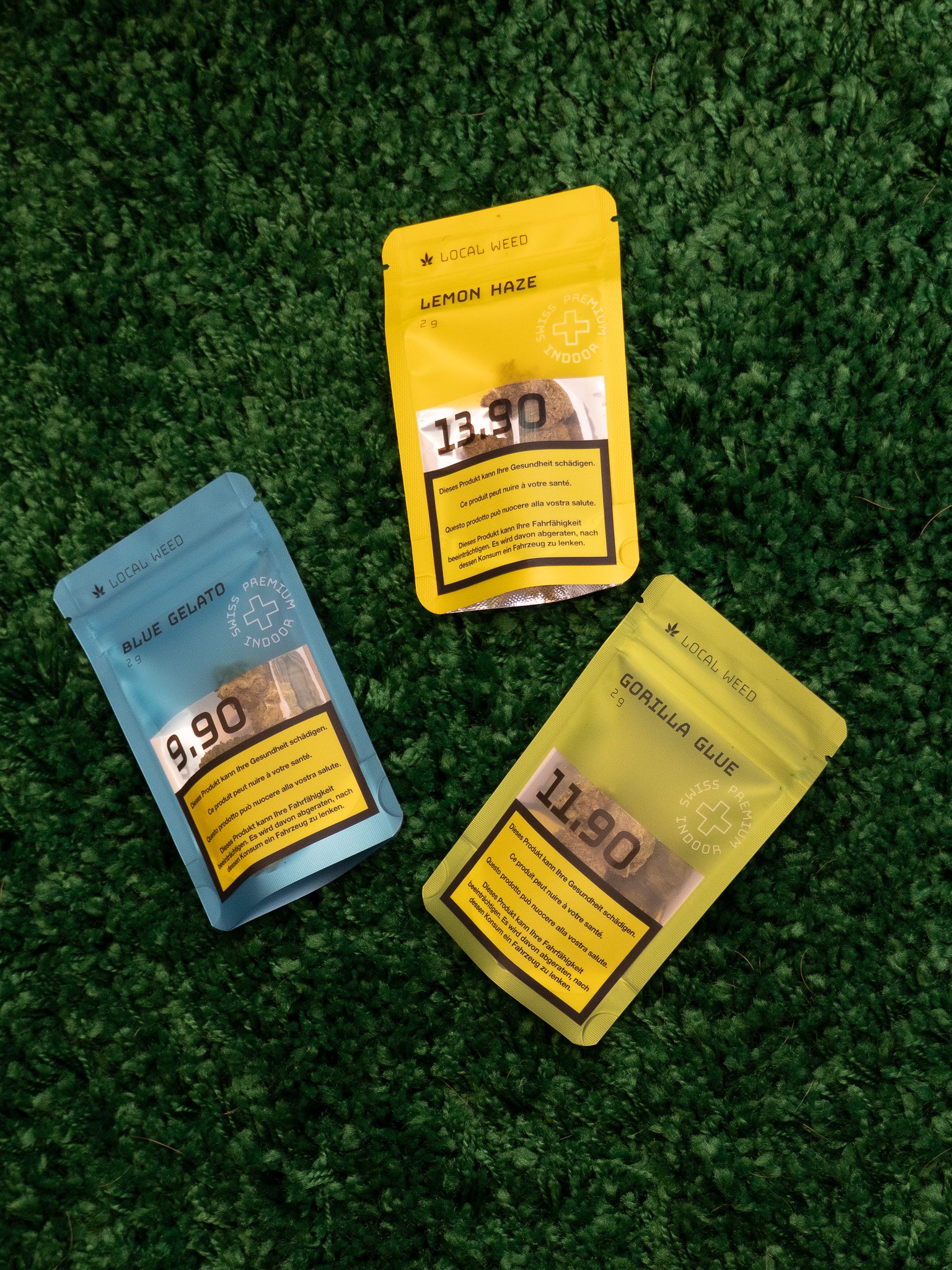Three colorful packaging pouches labeled with strain names and prices placed on a green leafy textured surface.