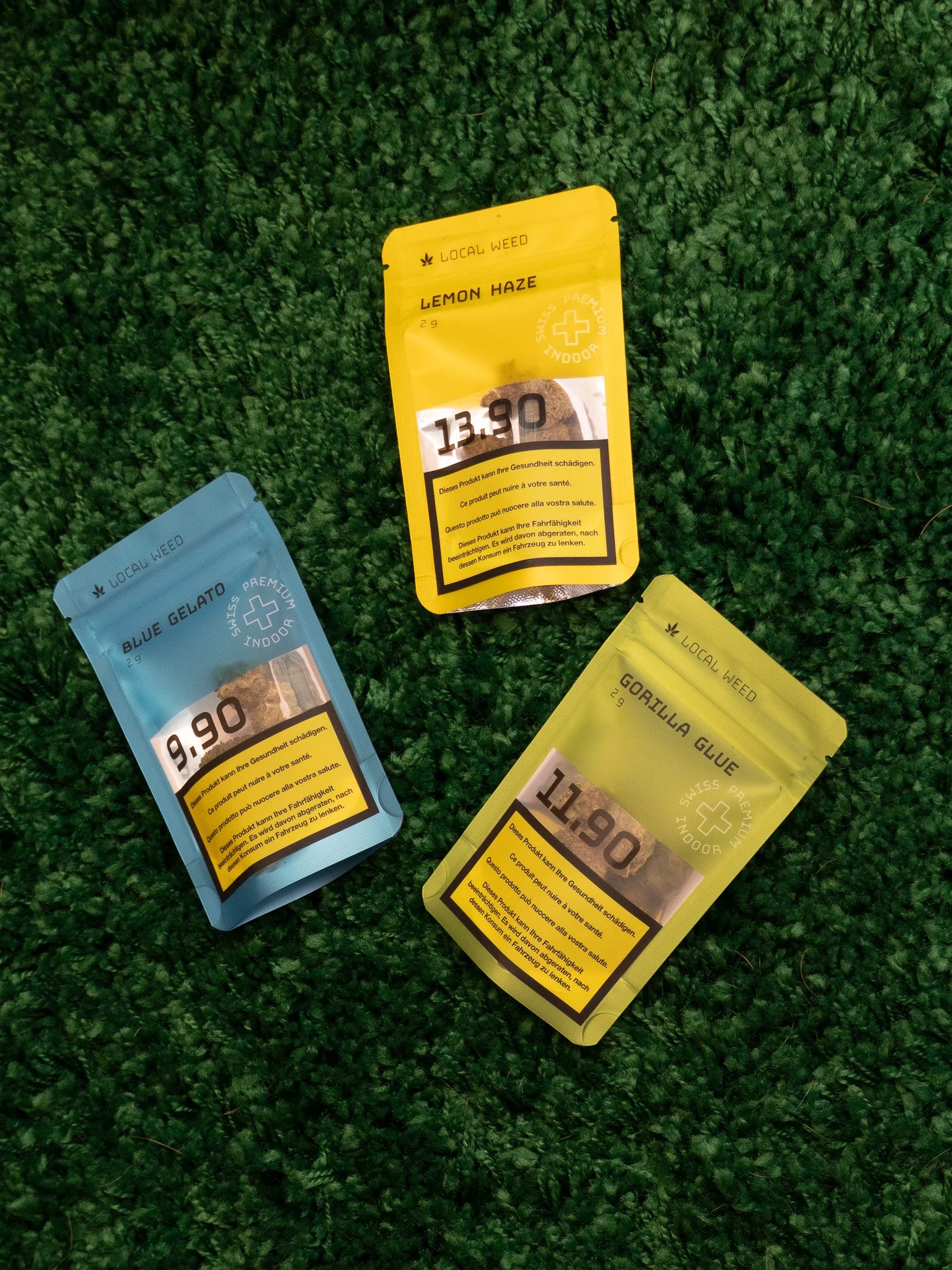 Four colorful packaging pouches of cannabis products on green grass. The pouches are blue, yellow, black, and yellow with the names Blue Gelato, Lemon Haze, Oregano Glue, and others. Each pouch has a cannabis-related label.